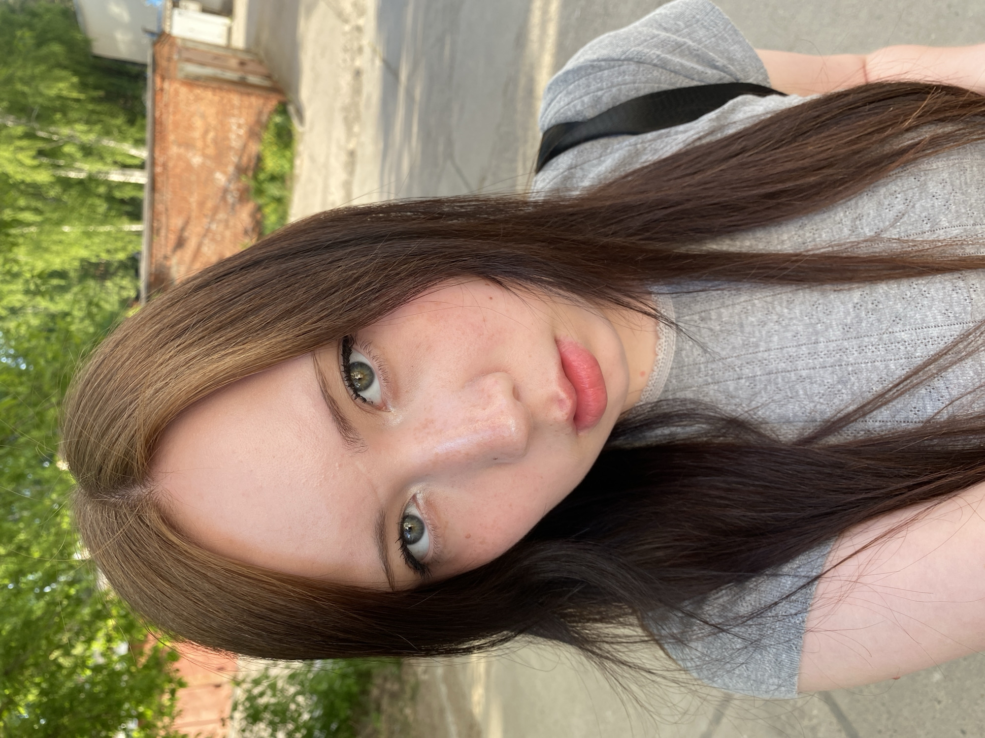 A young woman with light skin and brown hair is angled towards the camera. She has green eyes and is wearing light makeup. She is wearing a gray ribbed shirt. The background features a building made of red brick and green foliage. A concrete path is visible behind her. The picture appears to be taken outdoors.