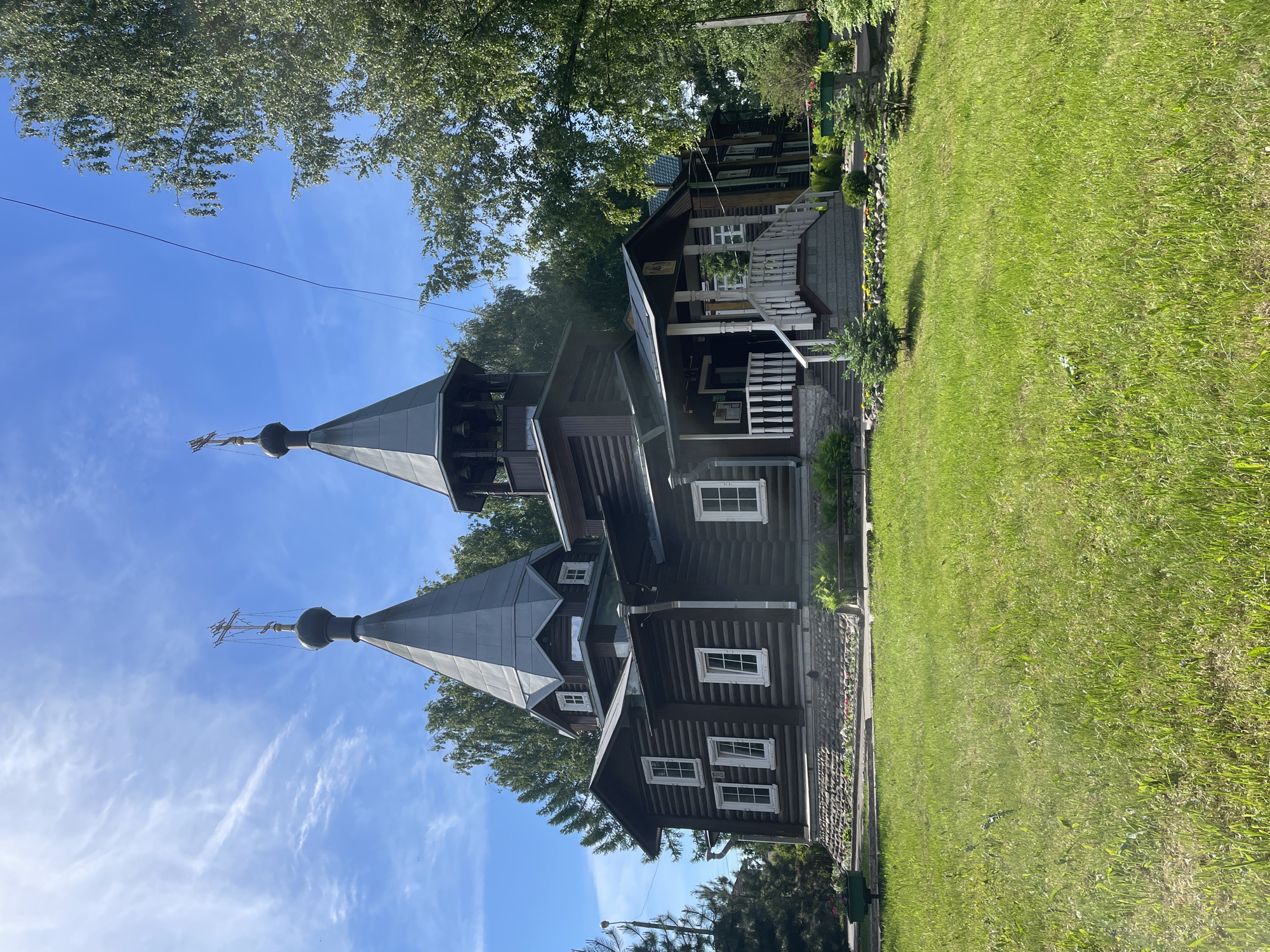 The image displays the Church of St. Andrew the First-Called, a wooden Orthodox structure situated at ul. Sergei Orlova, 15A in Vologda, Russia. The building is constructed from dark brown horizontal logs, resting on a base of irregularly shaped grey and brown stones. Two prominent, multi-tiered spires with grey metallic conical roofs rise from the church's main body. Each spire is capped with a small, dark, bulbous dome from which a tall, slender Russian Orthodox cross extends. The spire positioned towards the right side of the church's facade integrates an open bell tower section, revealing multiple dark-colored bells within.
The church features several white-framed rectangular windows, including two vertically stacked pairs on the left side of the main structure and individual windows on other facades. A covered porch, accessible via grey steps, extends from the front of the building on the right side, supported by white posts and featuring white wooden balustrades.
The foreground consists of an expansive, light green lawn with varied grass density. Immediately surrounding the church's foundation and porch steps, landscaping includes small evergreen shrubs, ornamental rocks, and patches of low-growing plants with visible red and yellow blossoms. The background is dominated by a variety of mature trees, including tall deciduous trees with light bark (consistent with birch) and conifers, suggesting a natural or park-like setting. The sky is bright blue with scattered, wispy white clouds. No individuals are present within the frame.