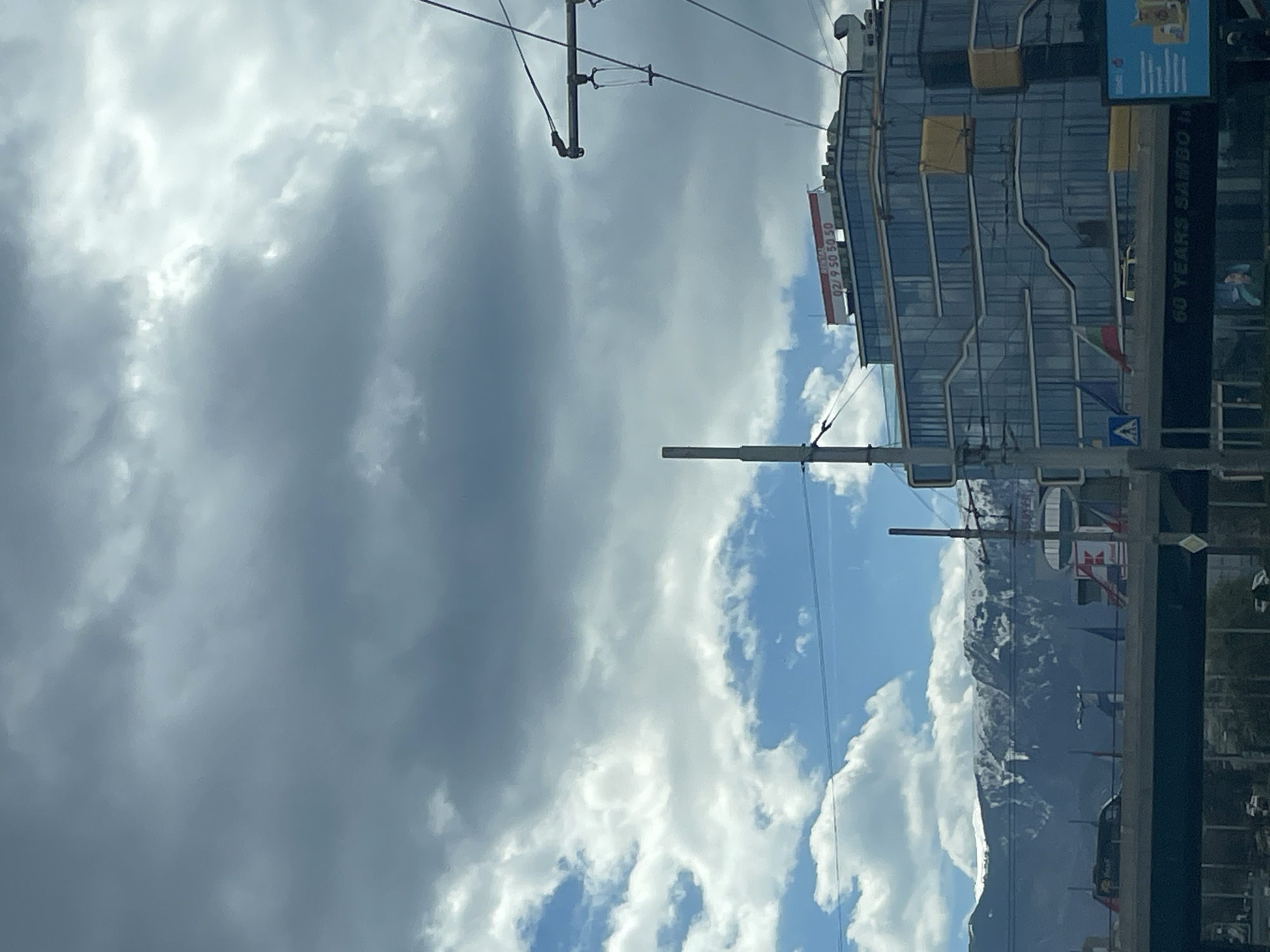 The image shows a street scene in Sofia, Bulgaria, with a view of a tall, modern building with multiple glass windows and a visible sign with the telephone number "02/9 50 50 50". A sign celebrating "60 Years SAMBO" is visible on the side of the building. Power lines and utility poles are in the foreground against a backdrop of a partly cloudy sky. In the distance, there is a mountain range with snow-capped peaks. The image appears to have been taken from inside a vehicle looking out.