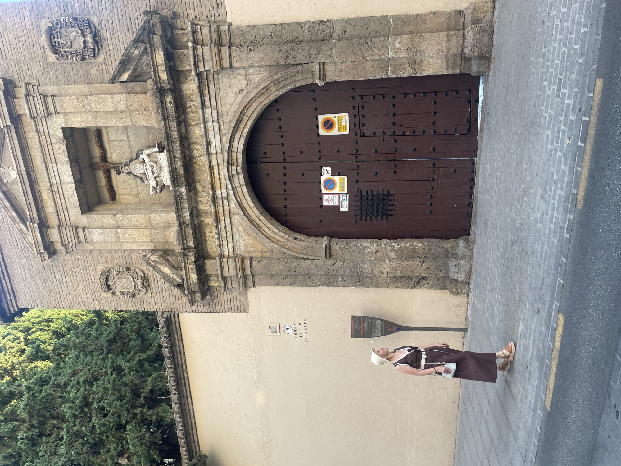 A woman, wearing a brown dress, white hat, and tan platform sandals, stands on a cobblestone sidewalk in front of a large, arched wooden door. The door is studded with dark metal rivets and has several signs attached to it, including two circular signs with a red diagonal line through them, indicating prohibitions, and a sign with a dog silhouette. Above the door, there is a niche containing a sculpture of a person holding a smaller figure, with a wooden cross behind them. Flanking the niche are stone carvings. To the left of the door, on a beige wall, is a sign with the number "9" and text that appears to read "PROHIBIDO FIJAR CARTELES." Adjacent to this sign is a tall, dark informational plaque. Behind the woman and the wall is a dense green tree. The scene is bathed in sunlight, casting shadows. The location is identified as Granada, Spain, specifically the entrance to the Capilla Real (Royal Chapel).