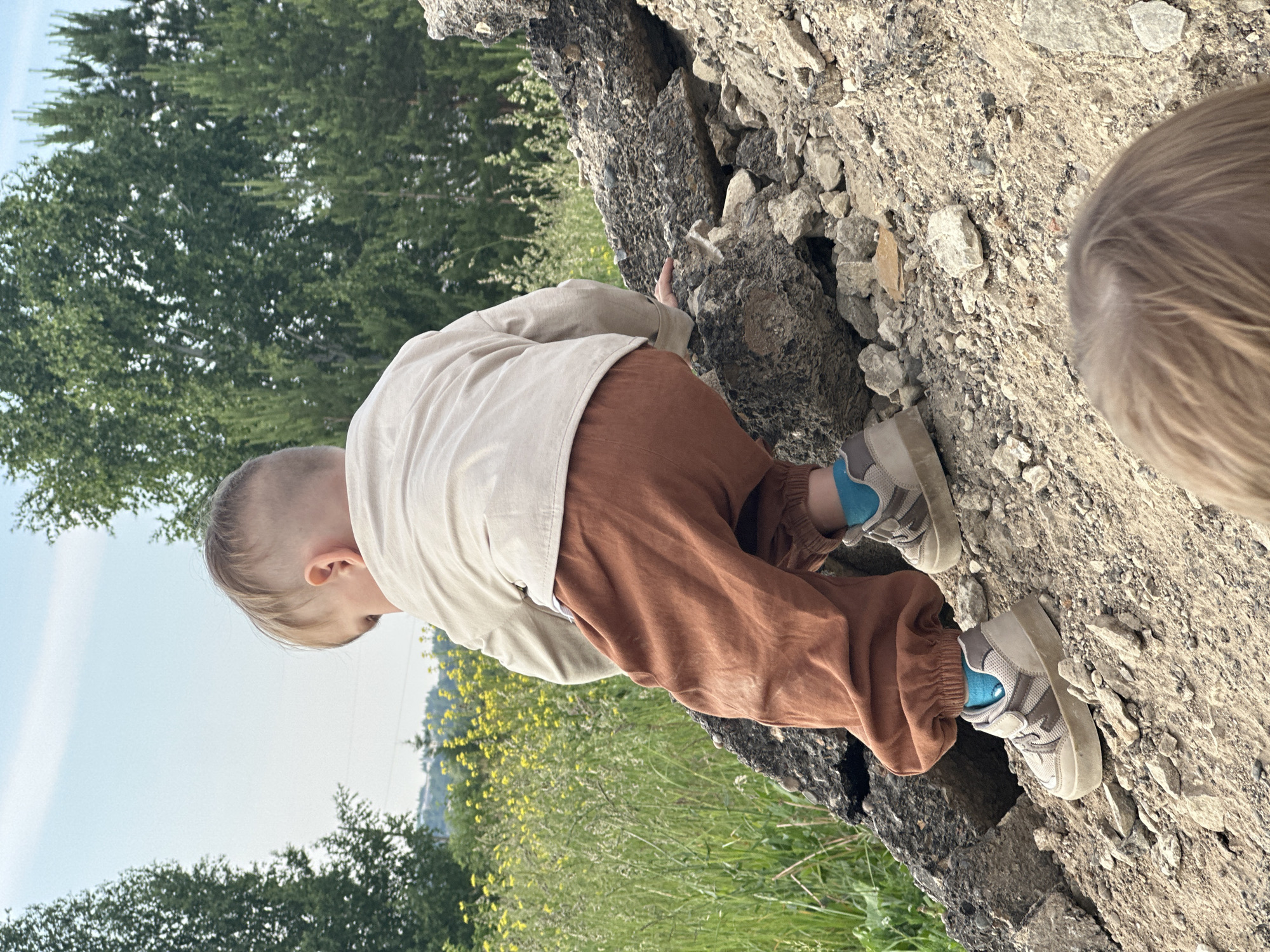 In a rural outdoor setting, likely on the outskirts of Estachi, Russia, a young child is actively climbing over a pile of rubble. The child, seen from behind and slightly to the side, wears a light-colored long-sleeved shirt, brown pants, blue socks, and beige sneakers. The rubble consists of large, irregularly shaped concrete and stone fragments. To the left of the rubble, a field of tall green grass dotted with yellow wildflowers stretches towards a distant treeline. Behind the field, a hillside with more trees is visible under a light blue sky. Another child's head, out of focus, is visible in the foreground. The overall scene suggests an unsupervised or semi-supervised play activity in a somewhat neglected or undeveloped area.