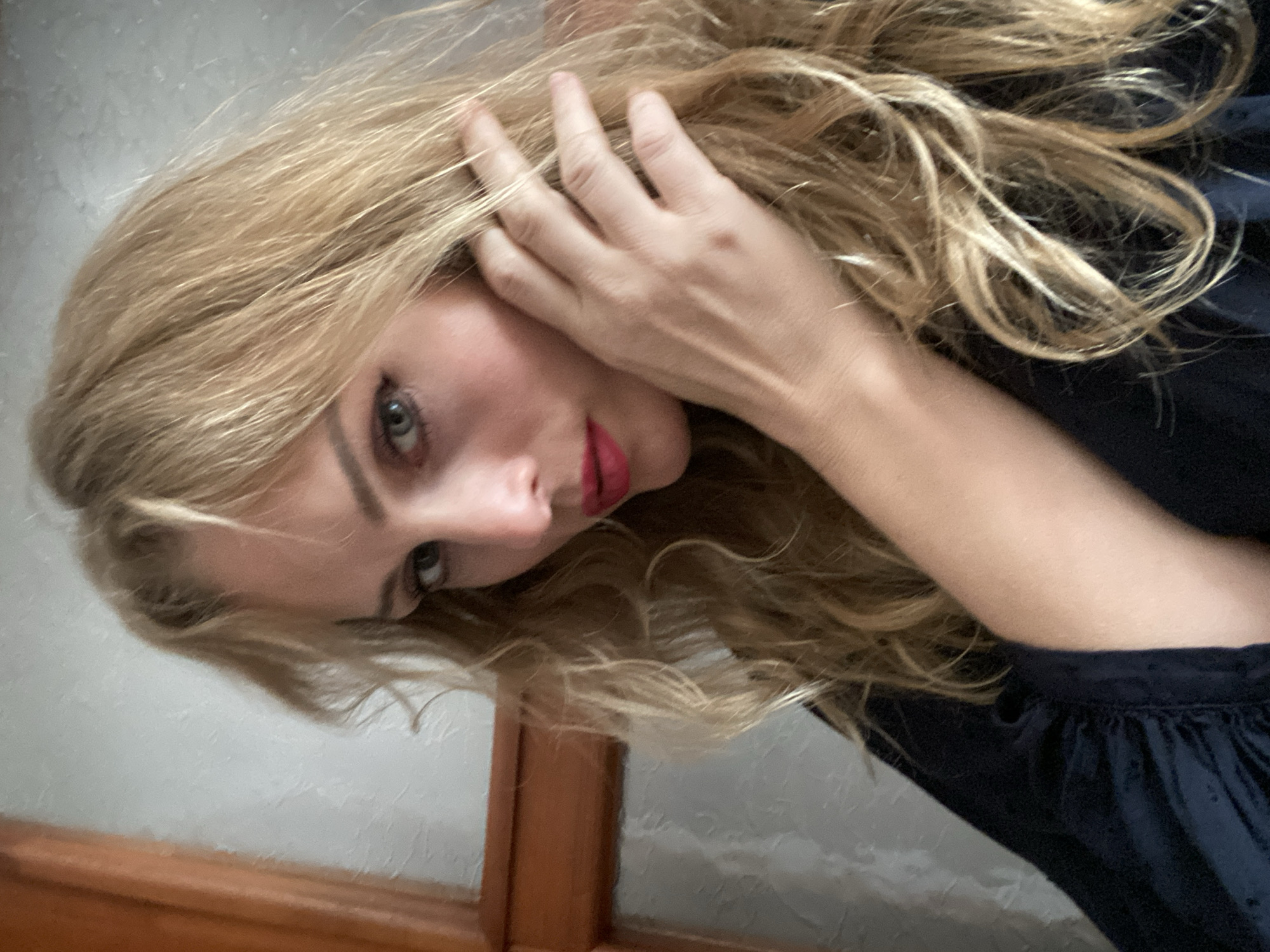 This is a close-up selfie of a blonde woman with her hand near her face and hair. She is wearing dark clothing and red lipstick. The background appears to be a wall and a wooden doorframe, suggestive of an indoor setting. There is no identifiable information within the image that would place it in the city of Lida, Belarus.