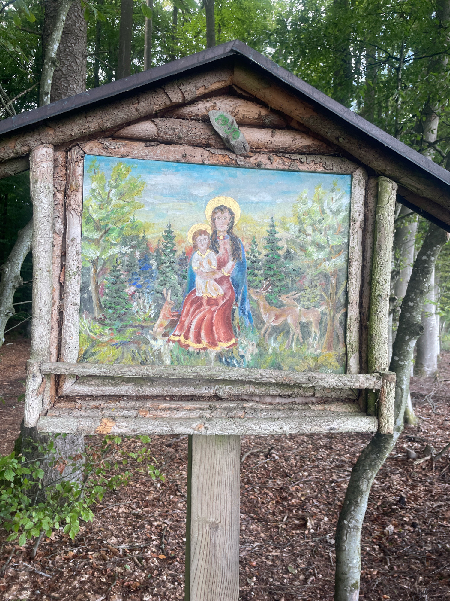The image shows a wooden structure, possibly a shrine or a birdhouse, set up in a forest. The structure has a pitched roof made of dark material, and the walls and supports are constructed from rough-hewn logs and branches. A painting is displayed prominently on the front of the structure. The painting depicts a woman in a red dress holding a child. They are surrounded by a forest scene with trees and deer. The painting has a naive or folk art style, with halos around the heads of the woman and child, suggesting a religious theme, likely depicting the Virgin Mary and Jesus. The surrounding environment is a forest floor covered in fallen leaves and twigs, with several trees visible in the background. One of the trees in the foreground has a distinctly curved trunk. The lighting suggests it is daytime, with dappled light filtering through the trees. The overall impression is a rustic, handmade shrine or artistic installation placed in a natural, wooded setting.