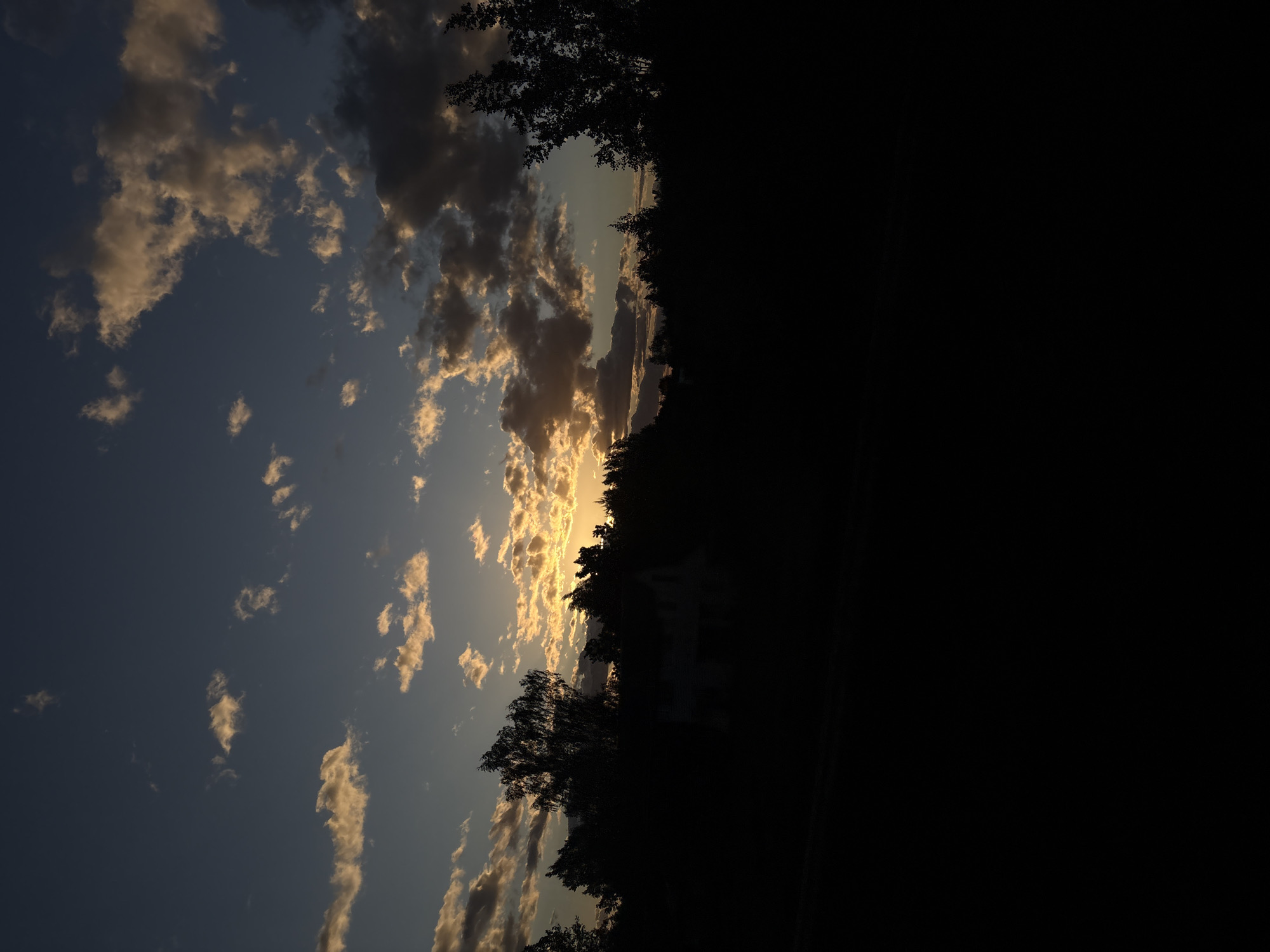 A vertically oriented photograph shows a sunset scene. To the left of the frame, a sky with blue hues is speckled with clouds that have their edges highlighted by the setting sun. The center features silhouetted trees and the suggestion of a structure or building, possibly a residence. To the right, the image is dominated by a field of black, indicating deep shadow or unlit space. The location is suspected to be somewhere in the Мажэйкаўскі сельскі Савет, Belarus. There are no observable people in this scene.