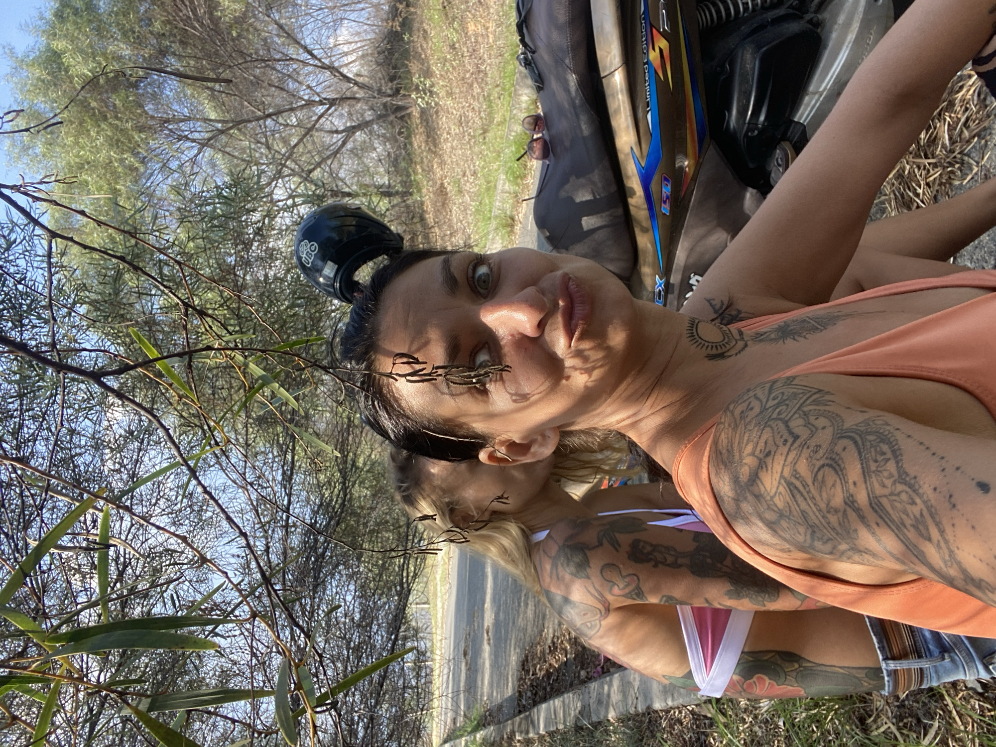 A close-up, low-angle, selfie-style shot captures two women in Phu Thuy Ward, Vietnam. The woman in the foreground, on the right, is wearing an orange tank top and has multiple tattoos on her left arm and shoulder. She is making a duck-face expression and has a black motorcycle helmet on her head, with one strap visible. Her hair is dark and pulled back.
Behind her, to the left, is another woman with blonde hair, wearing a pink and white bikini top. She has extensive tattoos on her arm and shoulder as well. She is leaning in towards the foreground woman, as if whispering in her ear.
The two women are seated next to a brown and gold motorcycle, with the brand "EX" and "150" visible on its side. Sunglasses are resting on the motorcycle seat.
The environment appears to be outdoors, with trees and foliage in the background, suggesting a rural or park-like setting. Sunlight filters through the branches, casting shadows on the scene. A paved path with a curb is visible to the left of the women. The ground is covered with dry leaves and grass. The overall lighting indicates it is daytime, likely in the morning or afternoon.