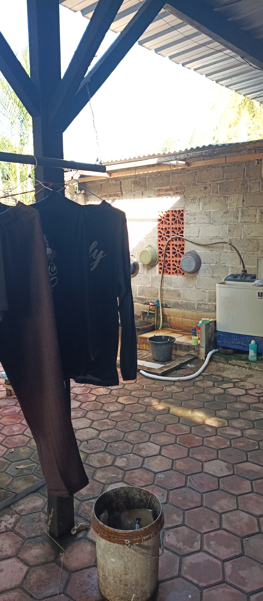 This image depicts an outdoor scene in Arjasa, Indonesia, likely a backyard or utility area. In the foreground, a patterned tiled floor is visible, with a weathered bucket containing some debris. Clothes, including a long-sleeved black shirt and brown trousers, are hanging on a clothesline under a wooden and corrugated metal overhang. In the background, a brick wall features a decorative ventilation opening. Various objects are present, including several buckets, a washing machine, and plumbing fixtures. The overall impression is one of a functional, albeit somewhat utilitarian, outdoor space.