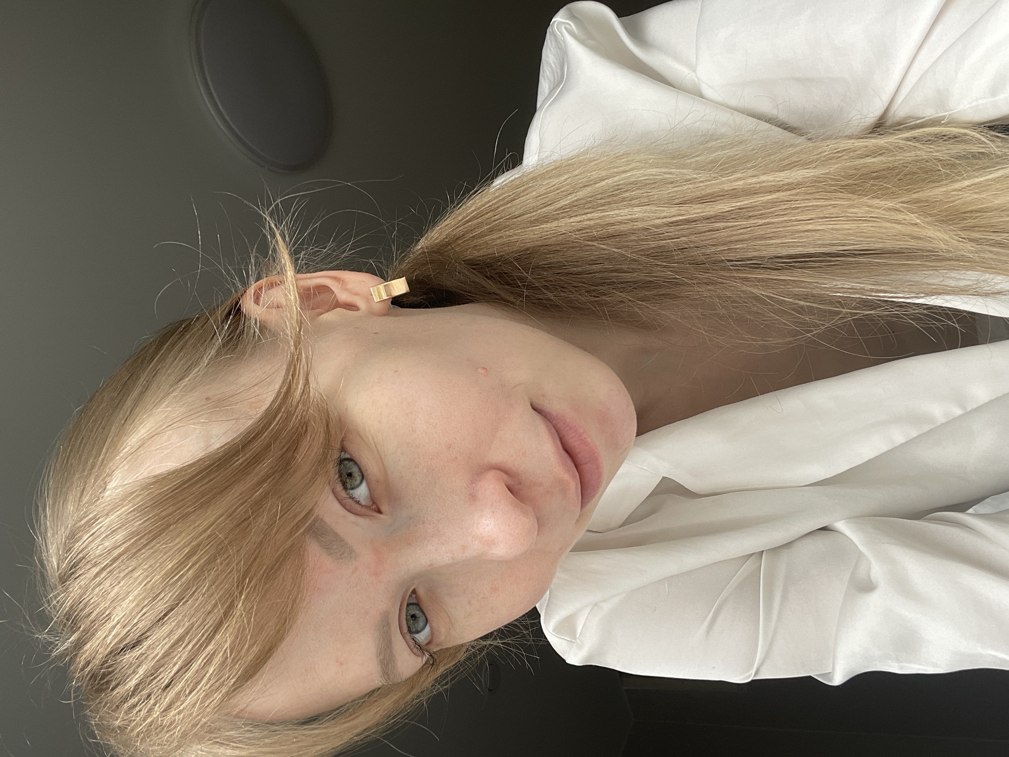A woman with long blonde hair is lying down, facing the camera. She has light skin and green eyes. She is wearing a white collared shirt. A gold rectangular earring is visible in her left ear. The background includes a gray wall with a circular fixture and dark furniture. The location cannot be determined as Perm, Russia, without further information.