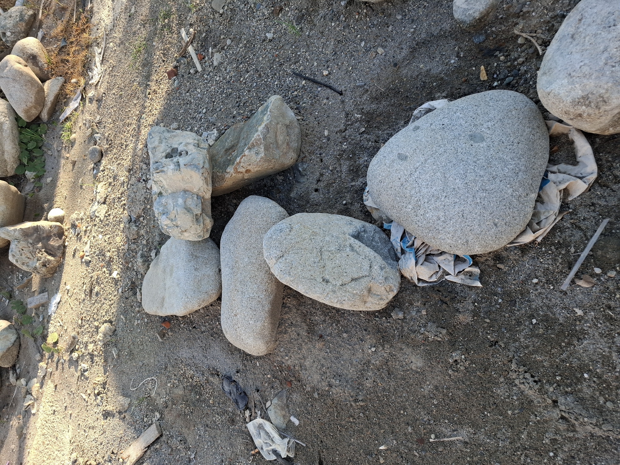 A high-angle shot captures a collection of rocks and debris on the ground in Gronggrong, Indonesia. The scene is dominated by various sizes of gray and tan rocks scattered across sandy soil. Some of the rocks are smooth and rounded, while others appear rough and irregular. Among the rocks, there are pieces of white and blue plastic bags, a small twig, and some dry, brown plant matter. The ground itself is a mixture of sand, gravel, and dirt, with patches of sunlight and shadow creating a textured surface. In the background, more rocks and some sparse green vegetation are visible. The overall impression is one of a natural, somewhat untidy outdoor environment.