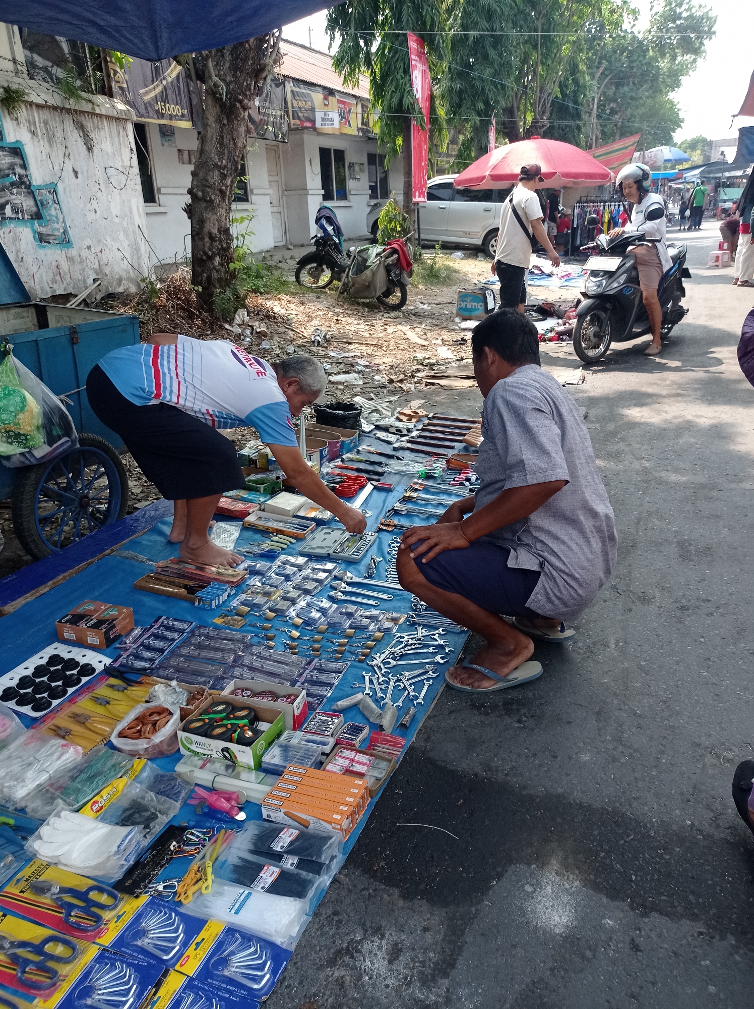 This image depicts a street market scene in Tegal, Indonesia, likely on a sunny day. Two men are engaged in commerce over a display of tools and hardware laid out on a blue tarp on the ground. The man on the left, wearing a blue and white striped shirt and black shorts, is barefoot and appears to be selecting an item from the display, leaning over it. The man on the right, wearing a grey shirt and dark blue shorts, is crouched down, observing the tools, and interacting with the seller.
The display includes a variety of tools such as wrenches, pliers, screwdrivers, locks, and what appear to be small tool kits. The items are neatly arranged in plastic packaging, boxes, and loose piles. In the background, other market stalls are visible, with awnings and umbrellas providing shade. Several motorcycles are parked nearby, and a few other individuals can be seen browsing or moving through the market. The environment suggests a busy, outdoor marketplace with a mix of permanent structures and temporary stalls. The ground is asphalt, with some areas appearing wet. A large tree with a thick trunk is prominent to the left of the frame, casting shadows. The overall impression is of a typical Indonesian street market where everyday goods, including tools, are sold.