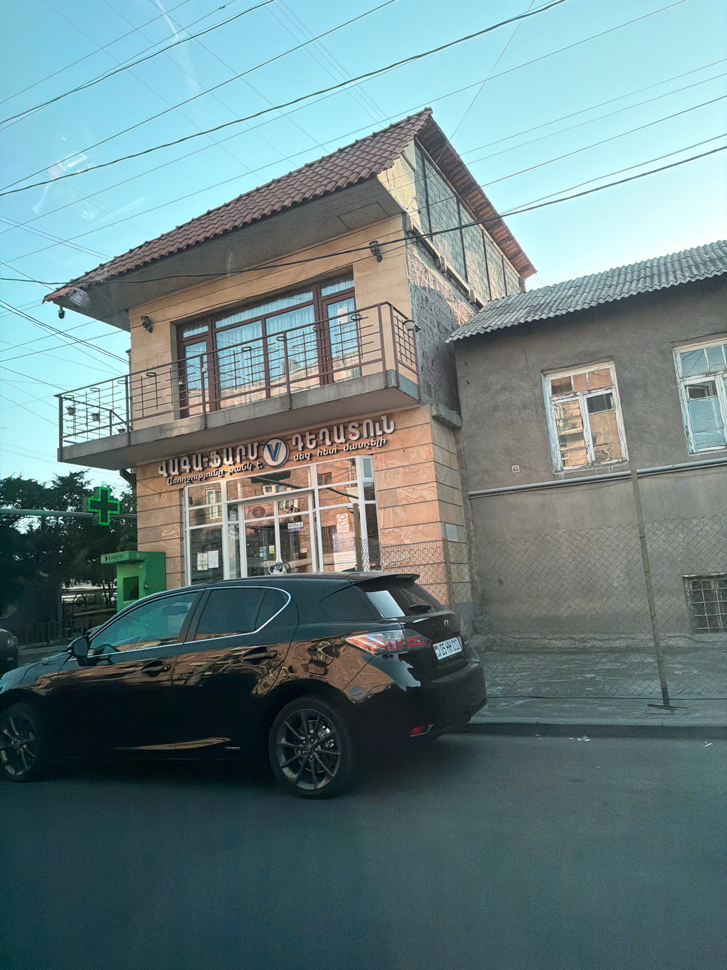The image depicts a street scene in Yerevan, Armenia, featuring a multi-story building with signage in Armenian. A black car is parked on the street in front of the building. The building has a modern facade on the lower floors with large windows and a balcony, while the upper story has a more traditional tiled roof. Adjacent to this building is a simpler, older structure with weathered walls and windows. A green cross sign, common for pharmacies, is visible on the left side of the frame, along with a green kiosk. Overhead, power lines crisscross the clear blue sky. The overall impression is a snapshot of a typical urban environment in Yerevan.