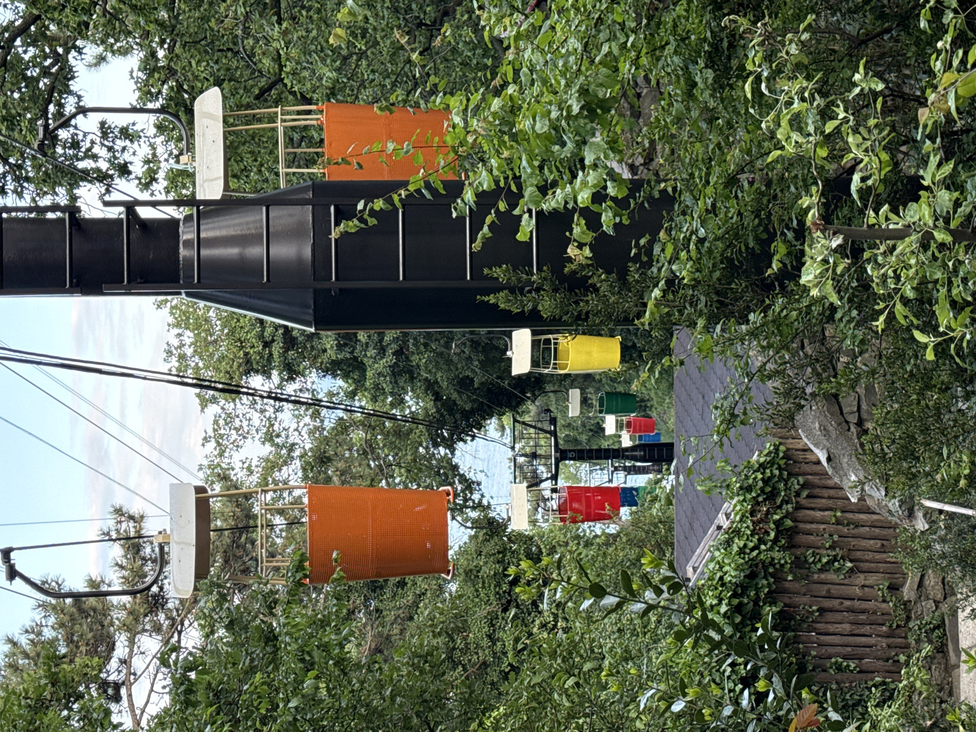 The image depicts a chairlift running through a heavily wooded area. The chairlift cars are painted in various colors, including white, orange, yellow, green, red, and blue. The chairs are suspended from cables that run between support structures. The support structures appear to be made of black metal. The surrounding environment consists of lush green foliage, with trees and bushes obscuring much of the background. The buildings of the Гаспринский поселковый совет, Ukraine area can be seen to the side, peeking through the forest canopy.