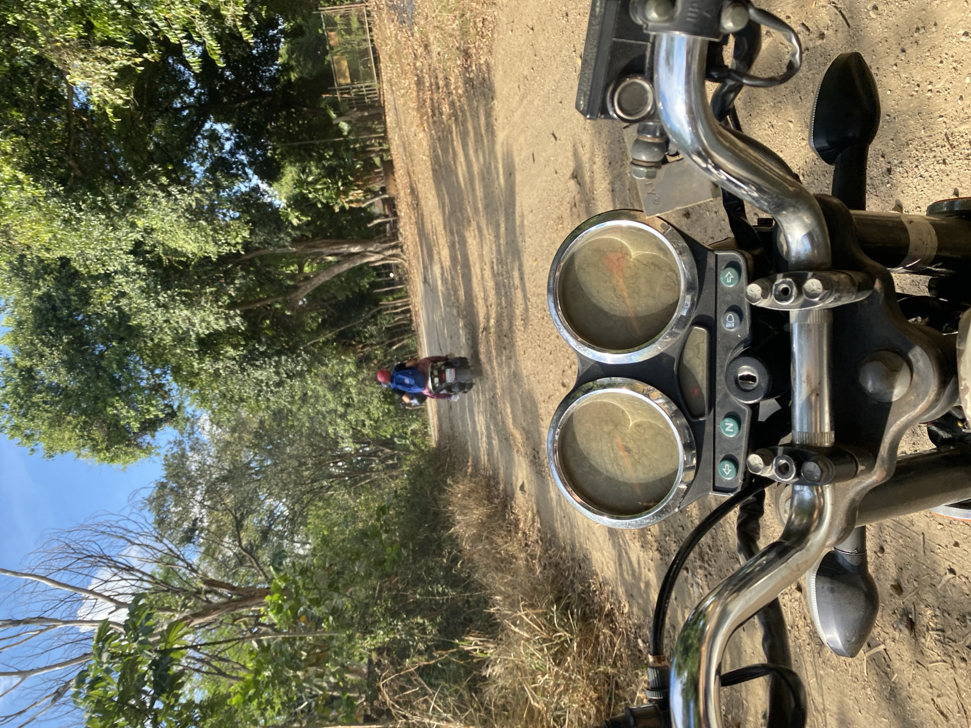 The image shows a view from the handlebars of a motorcycle on a dirt road. In the distance, another motorcycle with a rider wearing a blue shirt and a pink helmet is traveling down the same road. The location appears to be a wooded area, presumably within the city of Tinaquillo, Venezuela, based on the geographic association. The foreground consists of the motorcycle's dashboard, handlebars, and mirrors, while the background includes trees, the road, and the second motorcycle.