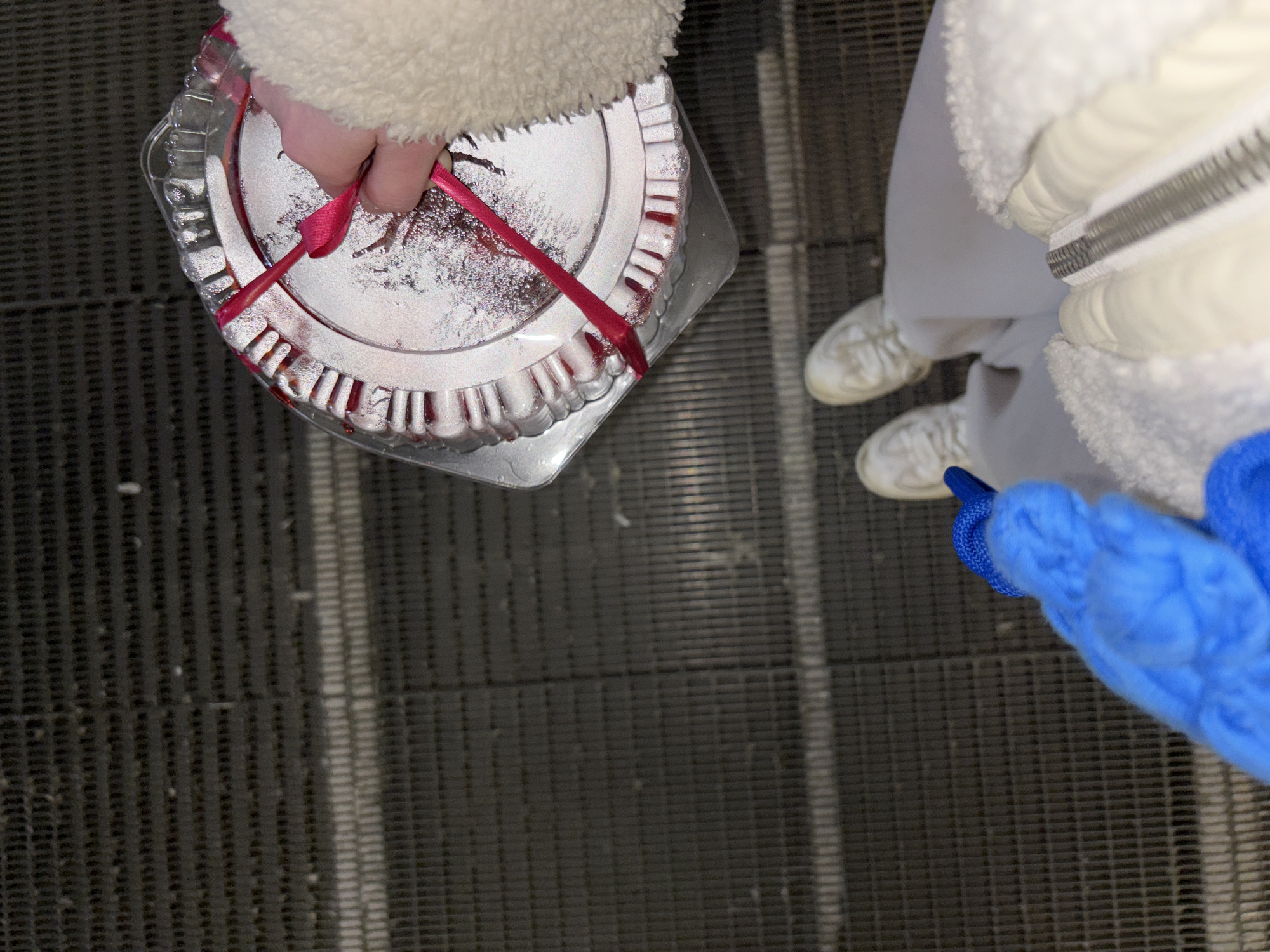 A top-down shot shows a person holding a cake in a clear plastic container with a red ribbon while standing on a moving walkway. The walkway has a dark, textured surface with parallel lines. Another person is partially visible with white shoes, white pants, and a white jacket. The second person also has a blue item, possibly a bag, visible in the lower right corner. The image appears to be taken indoors. There is no visual information to confirm the location as Perm, Russia.