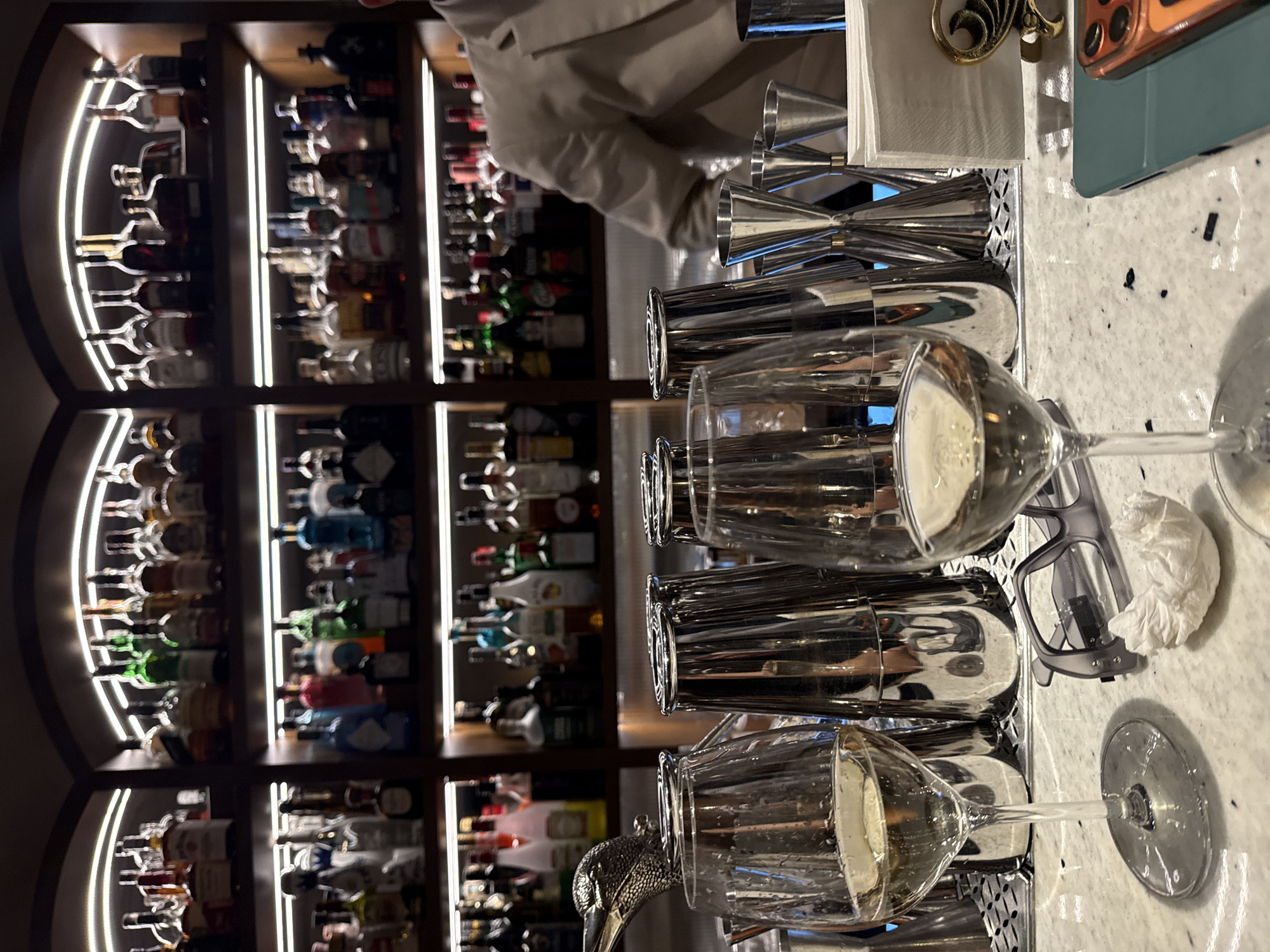 The photograph shows a bar setting. In the background, shelves are stocked with numerous bottles of liquor. In the foreground, glasses of sparkling wine are arranged on a bar counter. A person wearing a light-colored jacket is partially visible near the center of the image. The location is likely Yerevan, Armenia, though the image alone cannot confirm this.