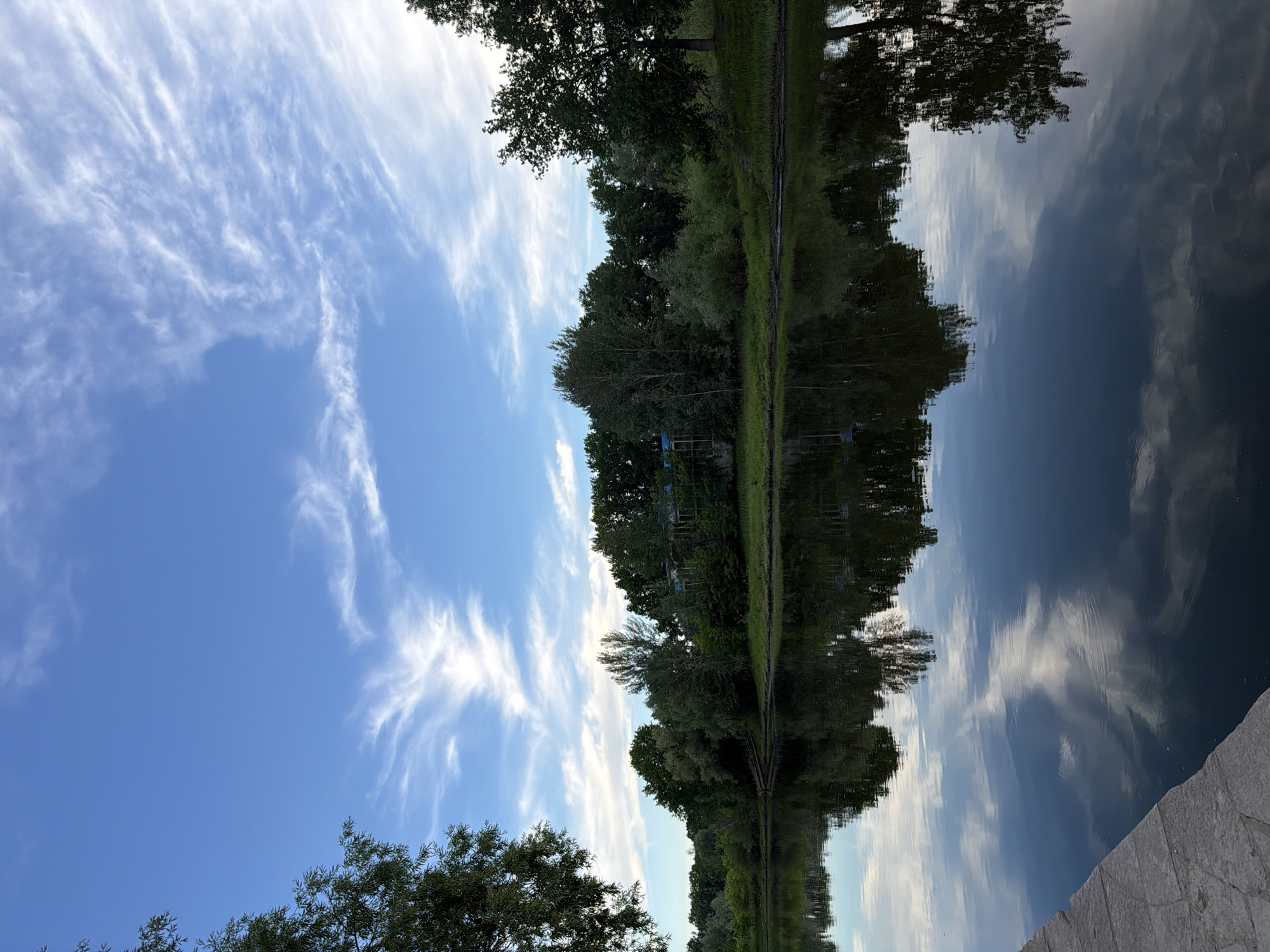 The image depicts a serene natural scene at the banks of the Volga River in Volzhsky, Russia. The foreground shows a stone embankment leading into the still water, which perfectly reflects the sky and the lush green trees lining the opposite bank. The sky is a vibrant blue, adorned with wispy white clouds that create a sense of depth and movement. A line of dense trees, varying in height and foliage, forms a picturesque backdrop against the sky. The reflections in the water are sharp and clear, mirroring the details of the trees and clouds. There are no people or animals visible in the frame, lending a peaceful and tranquil atmosphere to the entire composition. The overall impression is one of unspoiled natural beauty.