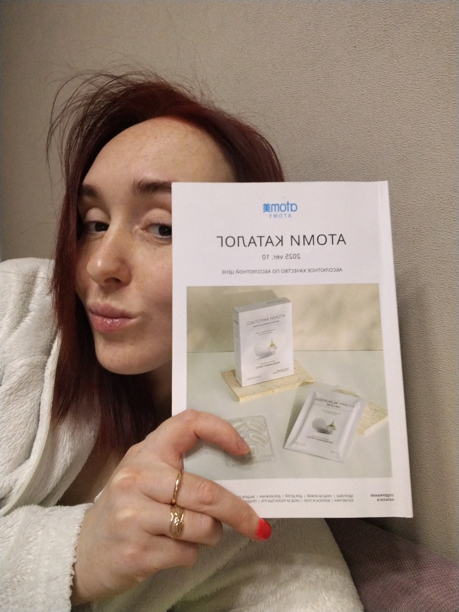 A fair-skinned woman with reddish-brown hair is posing indoors, holding a brochure in front of her. She is wearing a white, plush robe. She has freckles and is making a kissing face. She is wearing two gold-colored rings on the ring finger of her left hand. Her fingernails on the same hand are painted with a red polish.
The brochure features a brand name "YMOTA" and some text written in reverse Cyrillic. It also includes images of the product: what appears to be a patch and a white sachet. A location (Izhevsk, Russia) is not discernible from the image.