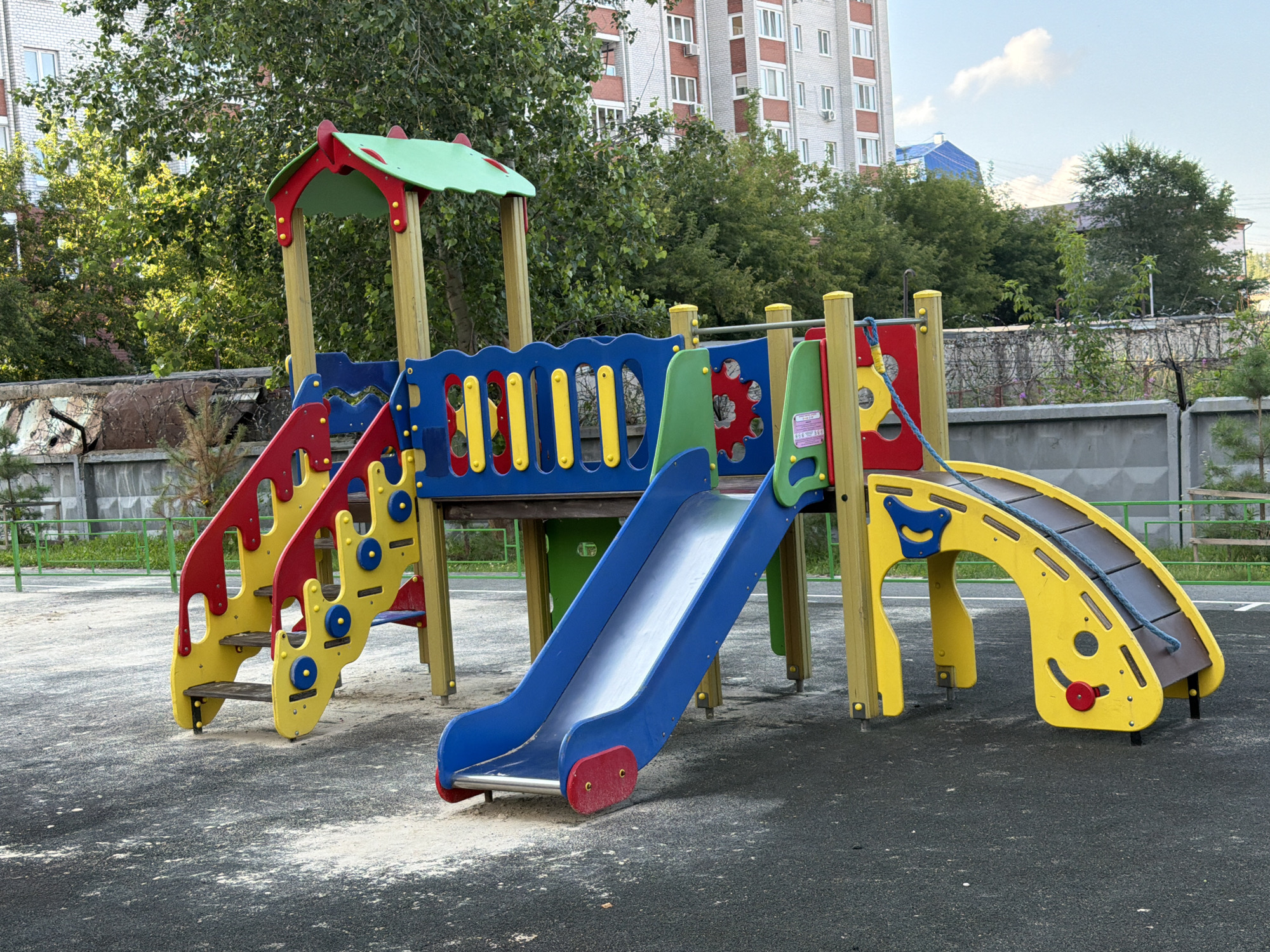 An empty, multi-colored children's playground structure, featuring slides, stairs, and platforms, is set against a backdrop of apartment buildings, trees, and a concrete fence topped with barbed wire. The ground surface is a mix of dark grey asphalt and scattered light-colored sand or fine gravel. The playground equipment is primarily yellow, blue, and red, with green accents on the roof of the main structure. A blue rope is draped over one of the yellow arched elements. The overall impression is one of a well-maintained but unused recreational area in an urban environment. The location is identified as Tyumen, Russia.