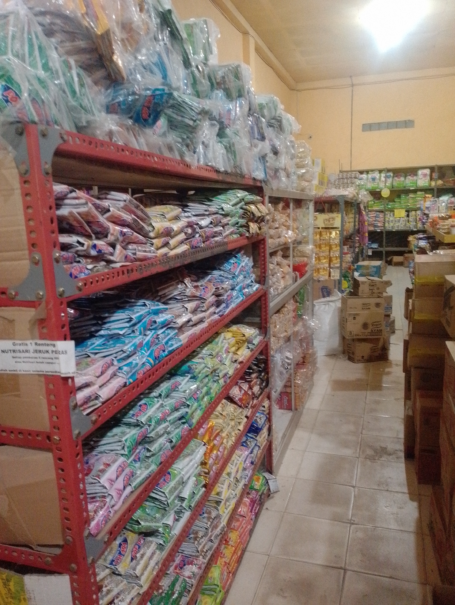 The image shows a retail store interior, possibly a minimarket or a small grocery store, located in the city of Hidayah Makmur, Indonesia. The scene is dominated by shelves stocked with various packaged goods, primarily snacks and beverages. On the left, a red metal shelving unit displays rows of brightly colored packaged items, many of which appear to be instant drink mixes or similar products. Some of these packages have text and imagery that is difficult to decipher due to the image quality and focus. A white label with Indonesian text is visible on the bottom left shelf, reading "Gratis 1 Renteng NUTRISARI JERUK PERAS".
Further into the store, more shelving units are visible, also laden with products. These shelves are a mix of metal and possibly wood. In the background, aisles are lined with more merchandise, including boxes and bags of food items. The flooring is tiled in a light, neutral color. The lighting in the store appears to be artificial, with some light fixtures visible on the ceiling, contributing to the somewhat dim and warm atmosphere of the establishment. There are no people visible in the image. The overall impression is one of a well-stocked, albeit somewhat cluttered, local retail environment.