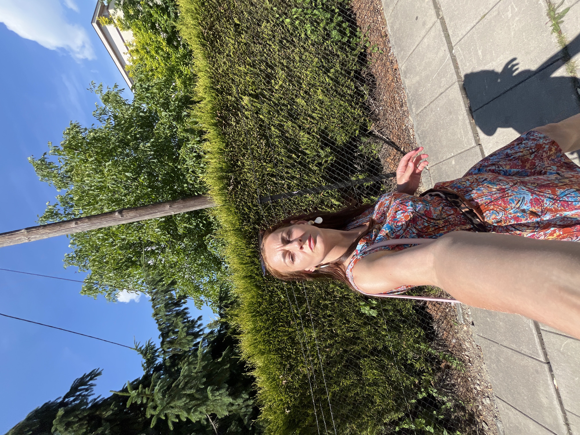 The image shows a person taking a selfie outdoors. The person, a woman with long brown hair, is wearing a colorful, patterned dress and large white earrings. She is looking towards the camera with her eyes closed and a slight smile on her face. She is leaning against a dense green hedge. Her right arm is extended, holding the camera. A shadow of her head and arm is cast on the concrete sidewalk next to the hedge. The sun is shining brightly, suggesting it is daytime and the weather is warm and pleasant. In the background, there are trees and a glimpse of a building. The scene appears to be in a residential or park-like setting.