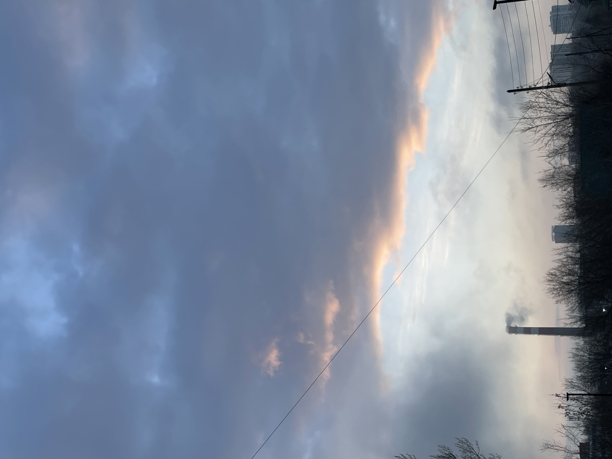 The image shows a view of the sky in Izhevsk, Russia, taken during twilight or dawn. Heavy, dark clouds dominate the left side of the frame, contrasting with lighter, brighter clouds tinged with orange and pink hues on the right, suggesting either sunrise or sunset. A power line diagonally crosses the scene. In the background, on the right side, a tall industrial chimney with smoke rising from it is visible, partially obscured by trees. Buildings can be seen in the far distance, partially obscured by the trees. The environment suggests an urban setting with industrial activity.