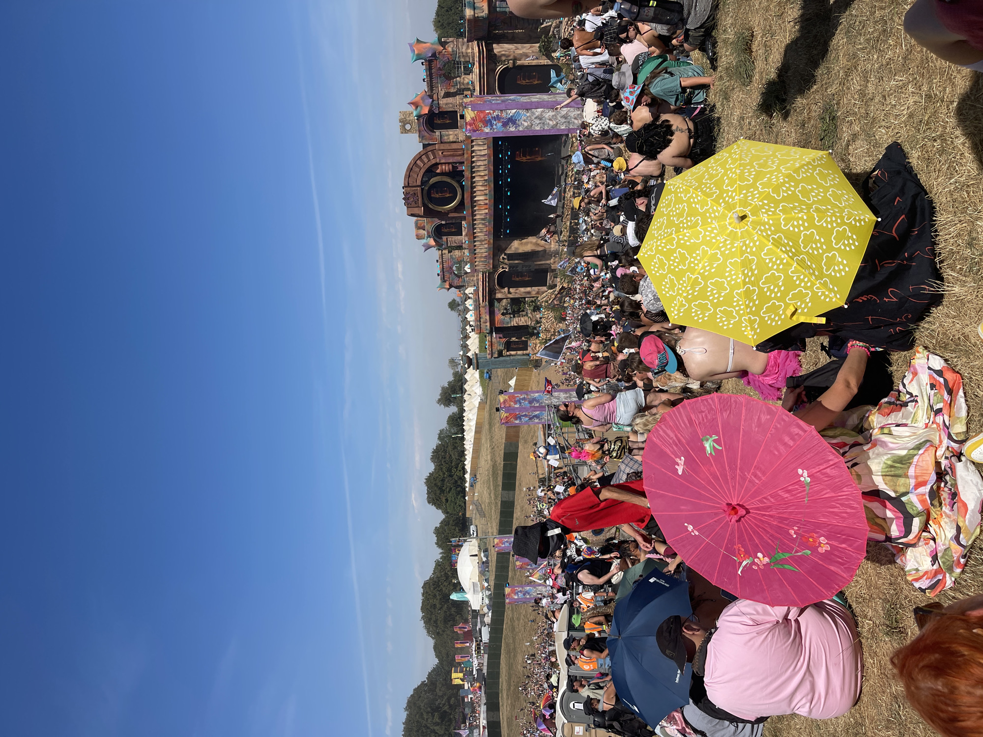 The image depicts a large outdoor gathering, likely a music festival, on a sunny day. A stage with an elaborate, ancient-ruin-like facade is visible in the background, with a crowd of people in front of it. The foreground shows a group of people sitting on dry grass, some using umbrellas for shade. Two prominent umbrellas, one bright pink with floral motifs and another yellow with a cloud pattern, are in the center of the foreground. The environment appears to be a grassy field with scattered trees and tents in the distance. The overall atmosphere suggests a vibrant event under a clear blue sky. There is no identifiable information about the city of Winchester, United Kingdom in this image.