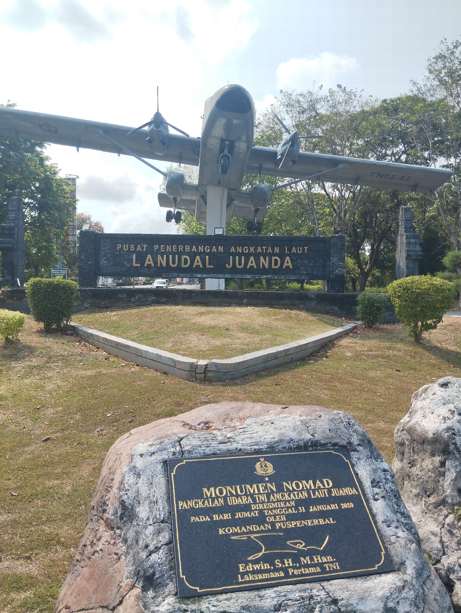 The image shows an outdoor monument featuring a decommissioned aircraft mounted on a pedestal. The aircraft is a twin-engine propeller plane, painted in a military gray color, with "TNIAL" visible on the wing. In front of the aircraft is a large, dark stone monument with gold lettering. The inscription on this main monument reads "PUSAT PENERBANGAN ANGKATAN LAUT LANUDAL JUANDA". Below this, in the foreground, is another monument set on a large rock. This smaller monument is a dark plaque with gold lettering that details the "MONUMEN NOMAD PANGKALAN UDARA TNI ANGKATAN LAUT JUANDA". It also states that it was inaugurated on Friday, January 31, 2020, by "KOMANDAN PUSPENERBAL", and signed by "Edwin, S.H., M.Han. Laksamana Pertama TNI". The surrounding environment consists of green grass, some dry leaves, scattered rocks, and trees in the background under a cloudy sky. The scene is likely at a military aviation base or a memorial park.