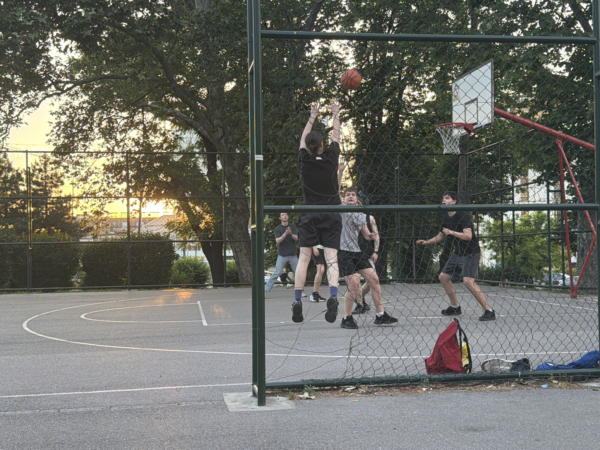 A daytime scene at a Belgrade, Serbia outdoor basketball court shows a group of young men playing basketball. One man is airborne, reaching up to shoot an orange basketball toward the hoop. Three other players are on the court, watching the play unfold. Two additional men stand by the sideline, also observing the game. The court is surrounded by a green chain-link fence. A red backpack and shoes are lying on the ground next to the fence. The background includes trees and buildings, with a sunset visible in the distance.