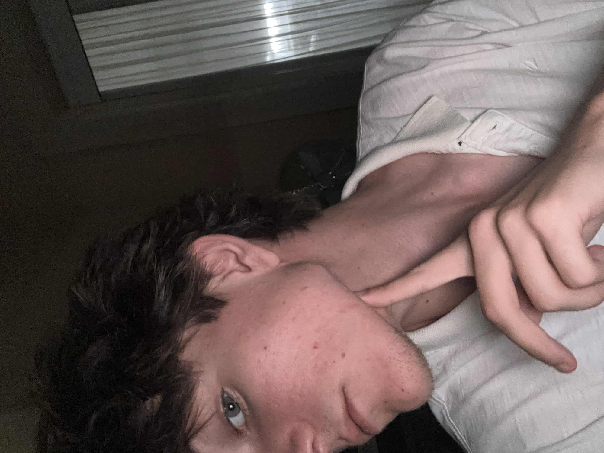 The image is a close-up, waist-up portrait of a young person with light skin and dark, messy hair, looking directly at the camera. They are wearing a white polo shirt. The person has their left hand raised, with their index finger touching their jawline. Their right hand is also visible, with fingers curled. The background is dark, with a partially visible sheer curtain on the right side, suggesting an indoor setting. The lighting is dim, with a strong light source coming from the right, creating shadows. The image does not contain any recognizable landmarks or features that would allow for its location to be identified as Cupra Marittima, Italy.