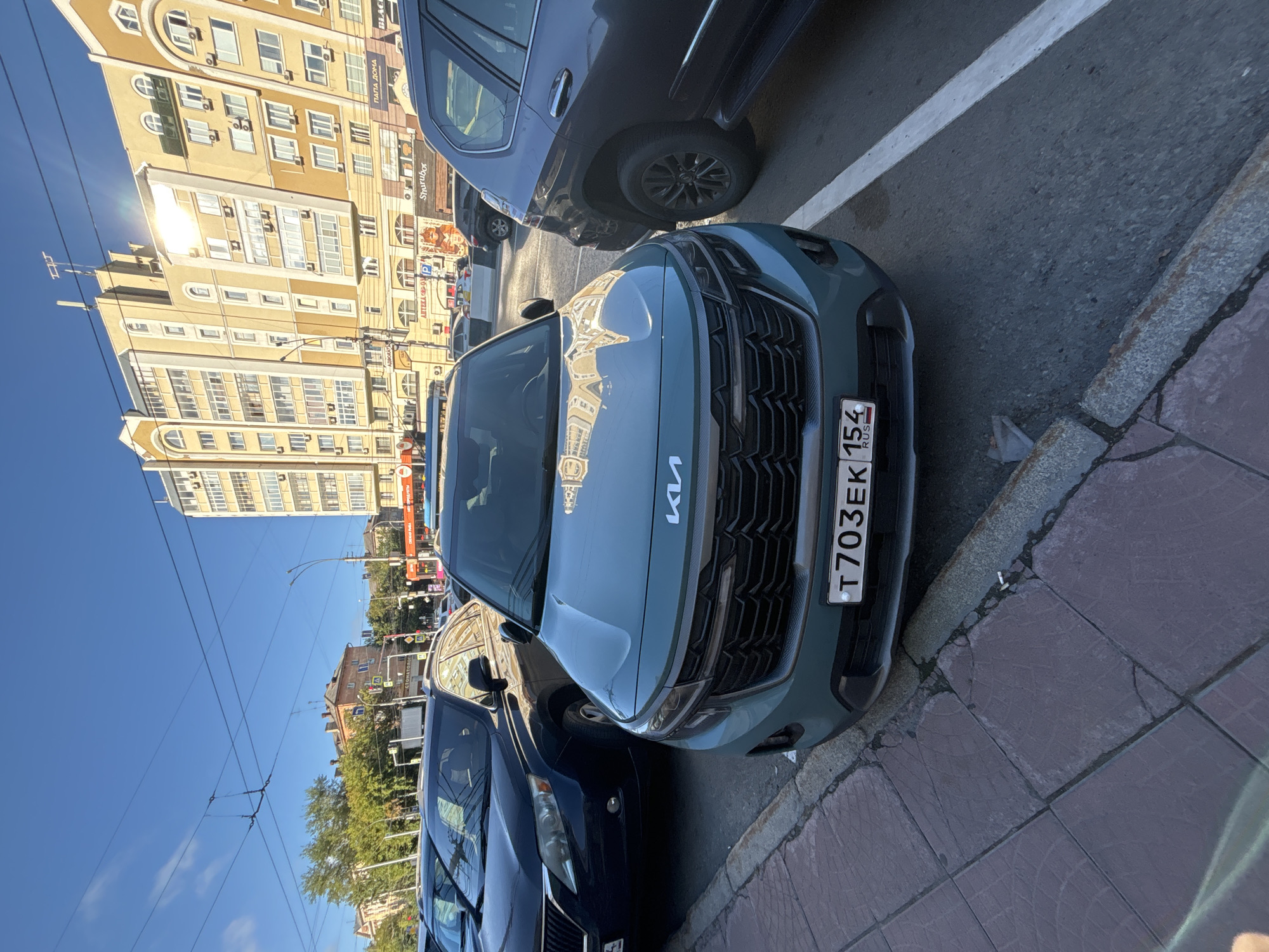 A teal Kia Seltos is parked on a city street in Novosibirsk, Russia, with its front bumper and license plate T703EK 154 visible. The car is positioned on the right side of the frame, angled slightly towards the viewer. Behind it, a dark gray SUV is also parked. In the background, a multi-story building with yellow and beige facade, featuring numerous windows and balconies, dominates the scene. A street with other parked cars and pedestrian walkways are visible in the mid-ground, along with traffic signals and overhead utility lines. The sky is clear and blue, with sunlight creating reflections on the building's surface. The sidewalk in the foreground is paved with gray square tiles and bordered by a concrete curb.