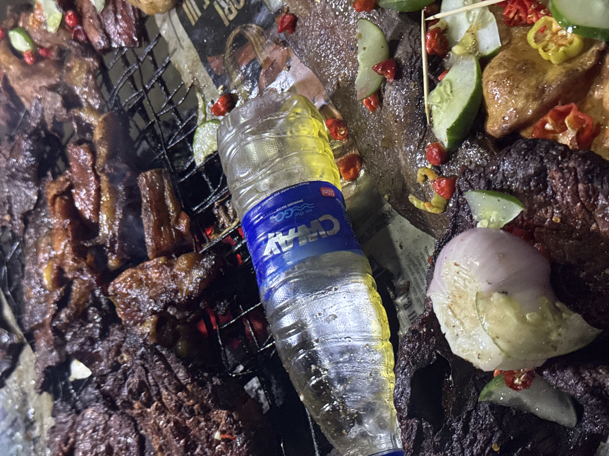 The image depicts a close-up view of grilled meat being prepared, with a plastic bottle of water lying on top of the food. The scene appears to be outdoors, likely at a food stall or market, given the presence of raw ingredients like onions, chili peppers, and sliced cucumbers scattered around the grilled meat. The meat itself is dark brown and appears to be well-cooked, with some pieces visibly charred. The water bottle, which has a blue label with white lettering, is partially filled with a yellowish liquid and has condensation on its surface, indicating it has been chilled. The overall impression is one of a casual, street-food setting. The image does not provide any specific clues to pinpoint the location in Abuja, Nigeria, beyond the general context of a food preparation environment.