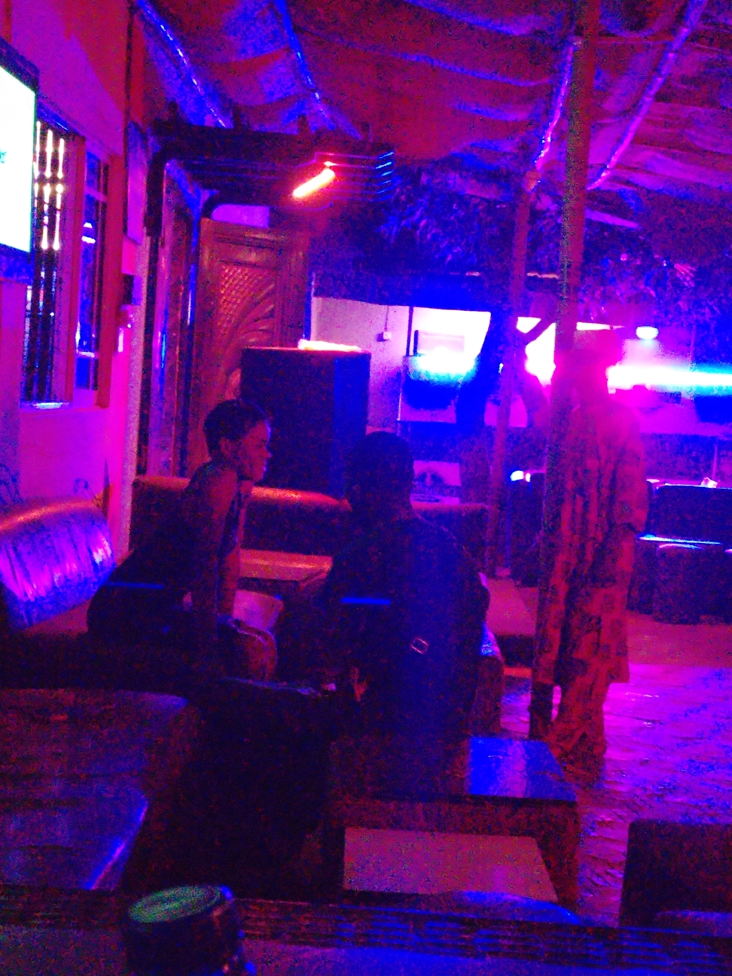 The image displays an indoor or semi-enclosed space, likely a lounge, bar, or club, under dim artificial lighting dominated by strong purple, blue, and red hues. The overall atmosphere is vibrant and hazy, suggesting a nighttime setting.
Three individuals are visible. On the left, a person with short hair is seated on a sofa, facing right, their profile faintly illuminated. They appear to be engaged with another person seated opposite them in the mid-ground, whose form is largely obscured by shadows and poor lighting. To the right, a third person stands upright, wearing a long, patterned garment, facing generally towards the left. Their facial features are not discernible.
The space is furnished with dark sofas or benches, some showing subtle patterns, and dark tables. The ceiling is draped with fabric or a similar material, supported by dark posts or pillars. A prominent horizontal red light fixture glows in the upper middle-left, while various blue strip lights are scattered throughout the background and foreground, enhancing the neon aesthetic. On the far left, a brightly lit rectangular area, possibly a screen or an internal window with vertical bars, adds to the light sources. In the immediate lower foreground, a dark circular object with a green top, possibly a bottle or can, rests on a table.
The scene captures a casual social gathering or a night out, with people relaxing and interacting in an environment characterized by its distinctive and colorful artificial illumination. No visible text is present in the image.