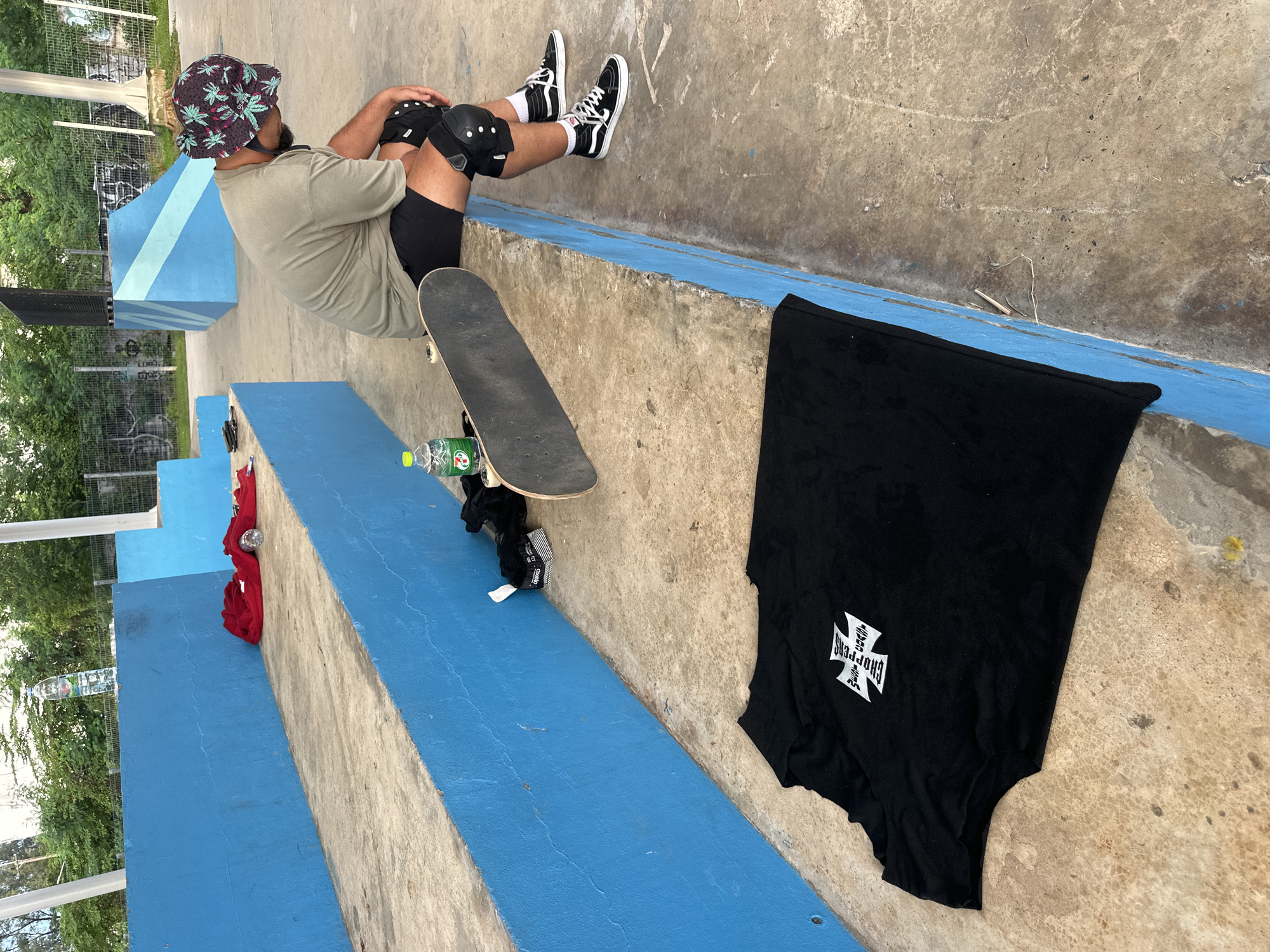 An individual wearing a green t-shirt, black shorts, knee pads, and a patterned bucket hat is sitting on a concrete ledge at a skate park. The individual is looking down and appears to be resting. A skateboard is placed on the ground next to the individual. A black t-shirt with a white cross logo is laid out on the concrete in the foreground. There are blue concrete structures and greenery in the background. The image is taken from a medium-wide shot, with the camera angled downwards. The location is implied to be Phuket City Municipality, Thailand.