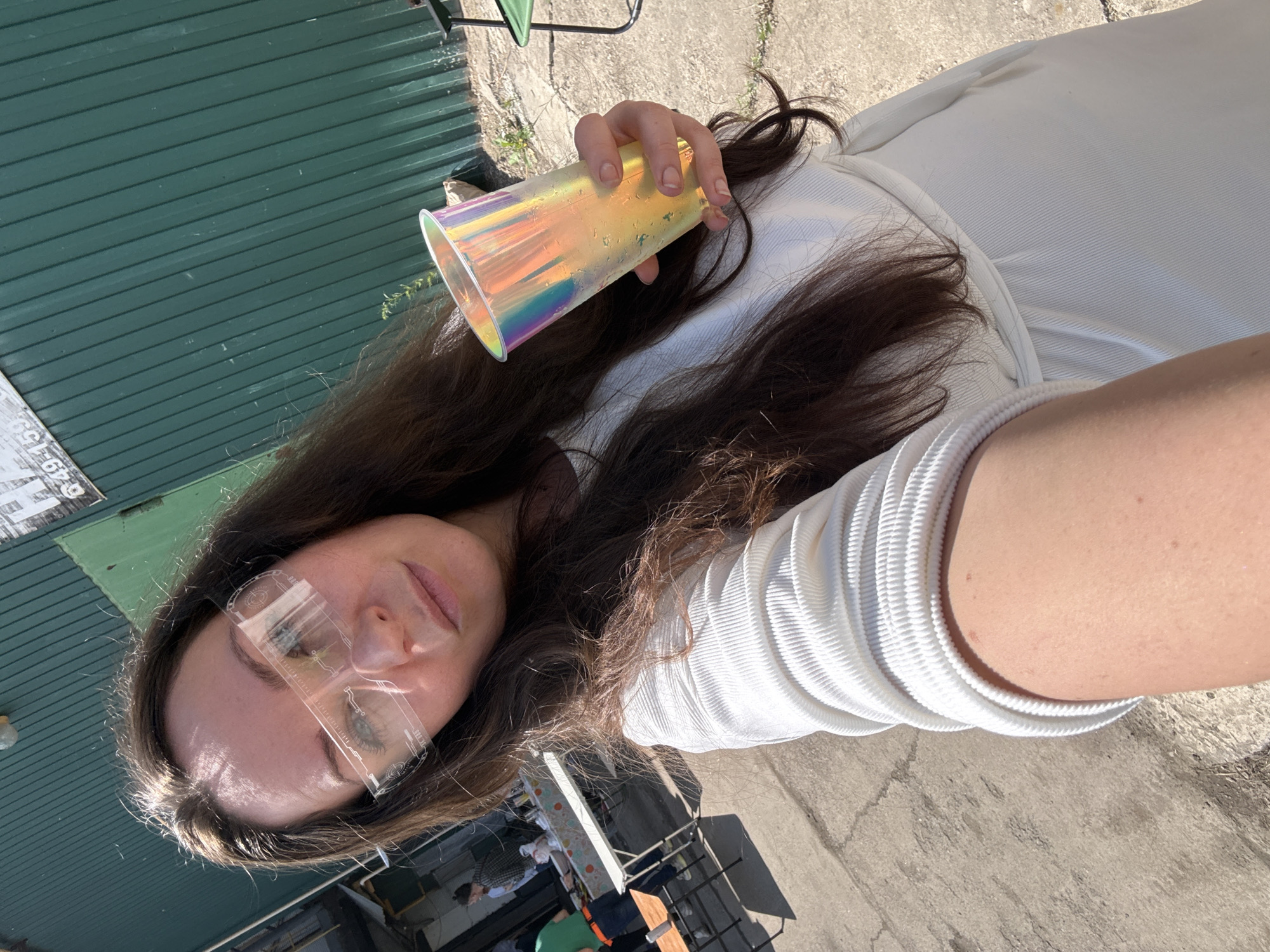 A woman is taking a selfie outdoors, holding a holographic cup in her right hand. She is wearing a white ribbed top and clear, futuristic-looking glasses. In the background, there's a green corrugated metal wall, a table with food, and two men. The ground is paved with concrete. The scene appears to be in a semi-industrial or outdoor event setting in Chaikovsky, Russia, given the style of the building and the casual gathering. The lighting suggests it is daytime.