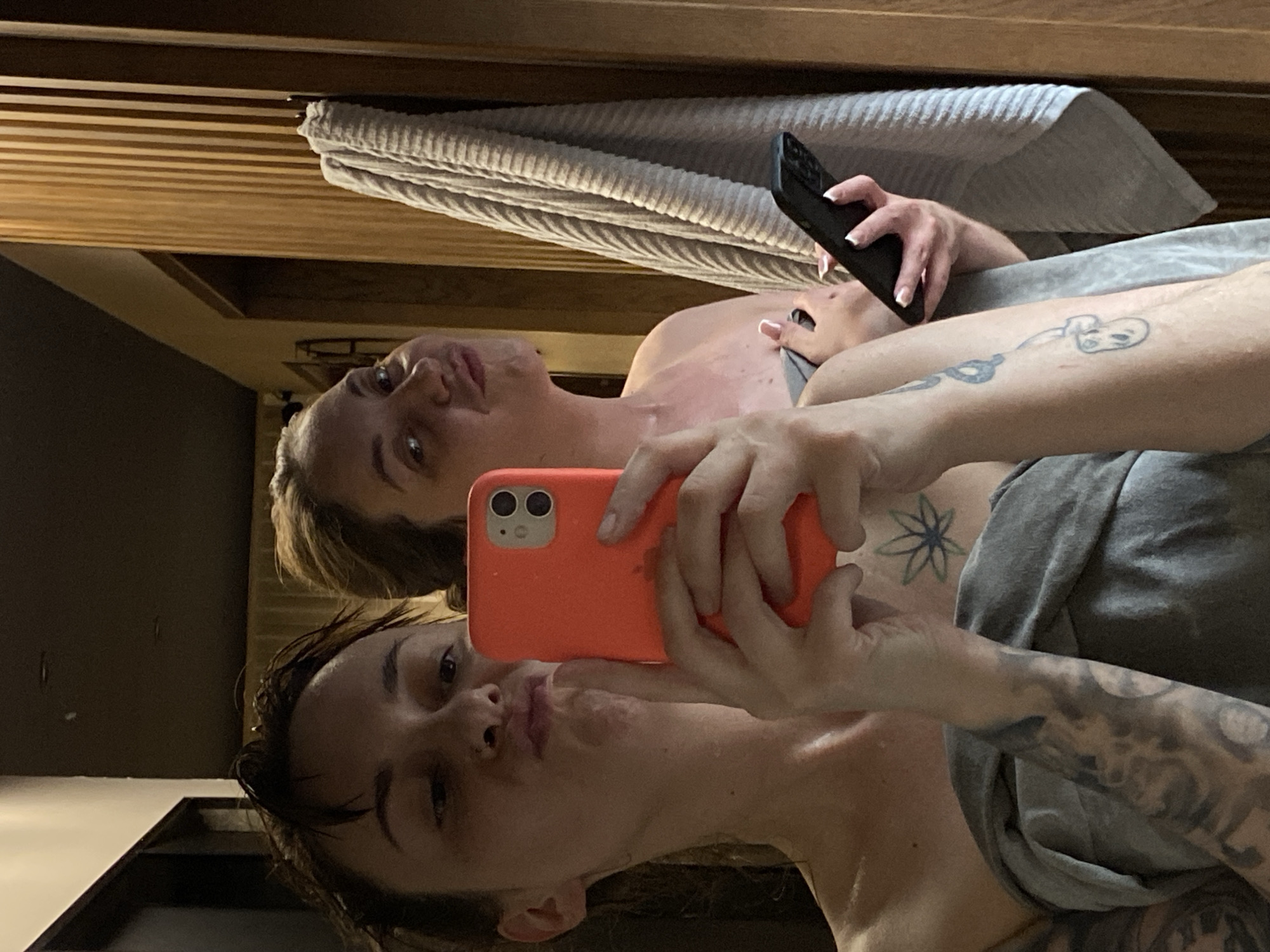 Two women are pictured in a selfie. The woman in the foreground is holding an orange iPhone, pouting slightly, with short dark hair. Behind her, the second woman is looking up, holding a black phone. Both women are partially visible, with tattoos visible on the woman in the background. The backdrop includes a textured wall, potentially wood paneling, and a grey towel or blanket. The location is unspecified and the city of Votkinsk, Russia, is not apparent from the image.