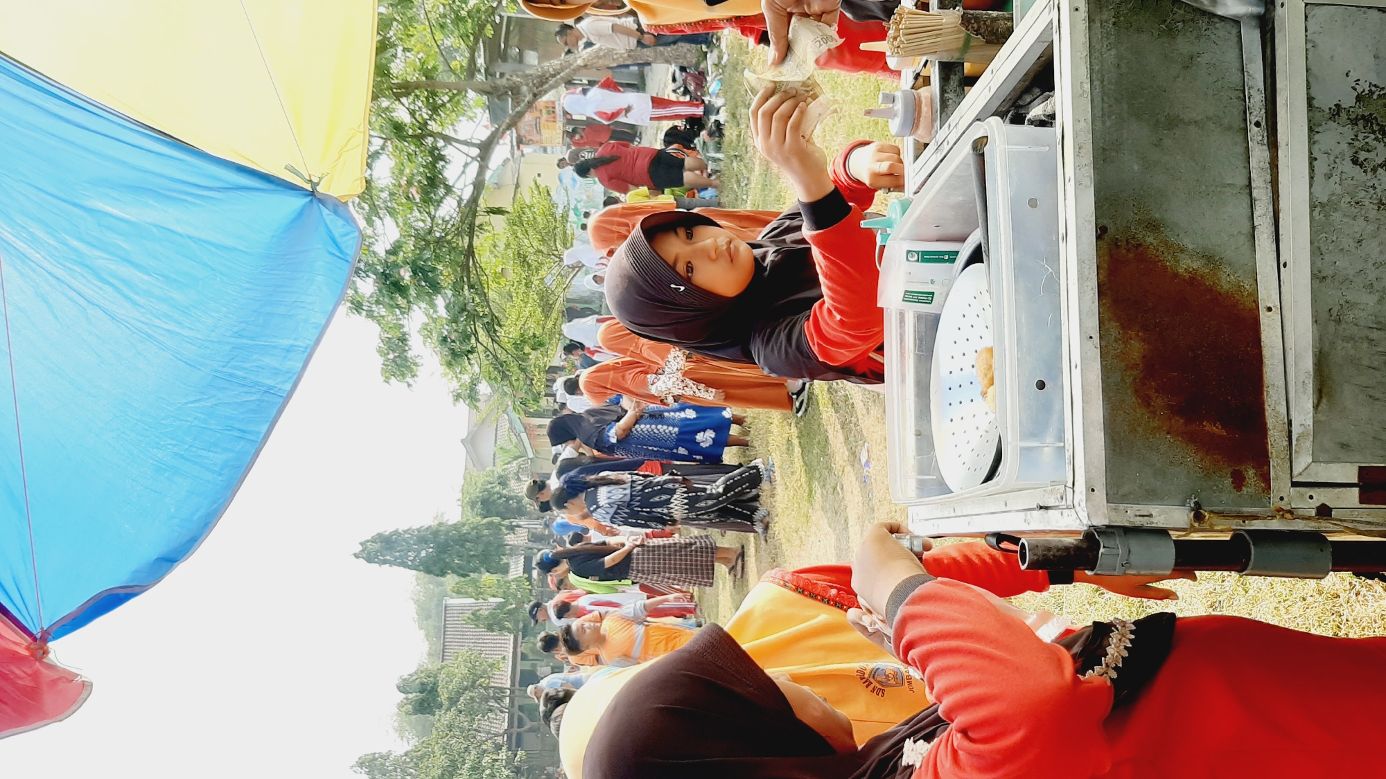 In Sumberjo, Indonesia, a group of people are gathered outdoors under a large blue and yellow tarp, suggesting an event or market. In the foreground, a young girl wearing a black hijab and a red long-sleeved shirt is handing money to another person who is partially visible. She appears to be purchasing food from a cart. The cart is made of metal and has a container with a white strainer and some fried food inside. There are also bottles of sauce and utensils on the cart. Other children and adults are milling around in the background, some wearing traditional Indonesian attire. The environment appears to be a grassy area with trees and buildings visible in the distance, indicating an outdoor setting within a community.