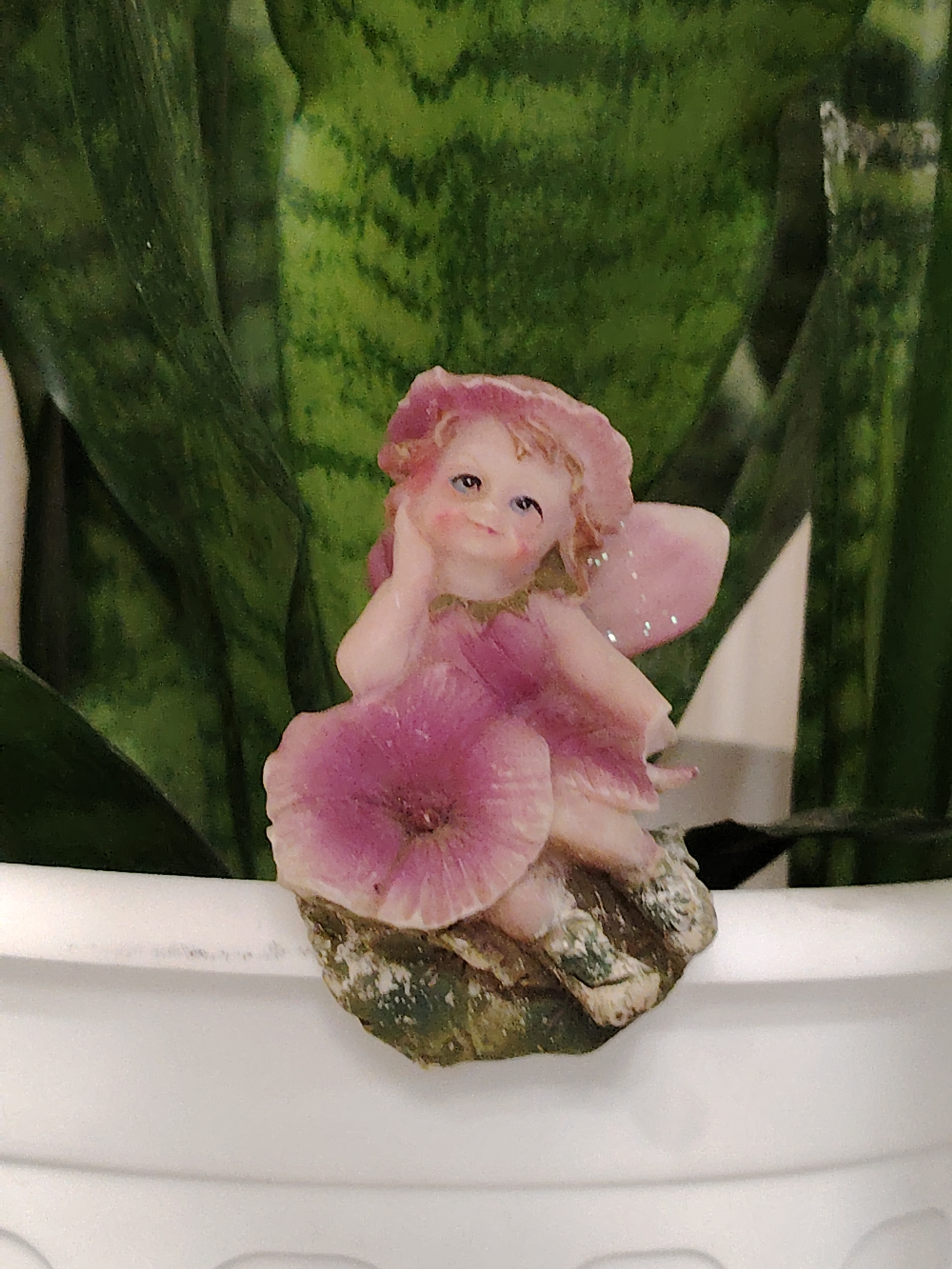 A close-up shot of a small, fairy figurine perched on the edge of a white, ridged flowerpot. The figurine is predominantly pink, with a flower-like hat, dress, and wings that have a glittery sheen. It sits atop a small, mossy-looking rock or base. The background is filled with tall, green, variegated leaves. The location information about Noviy, Russia, does not apply to the image.