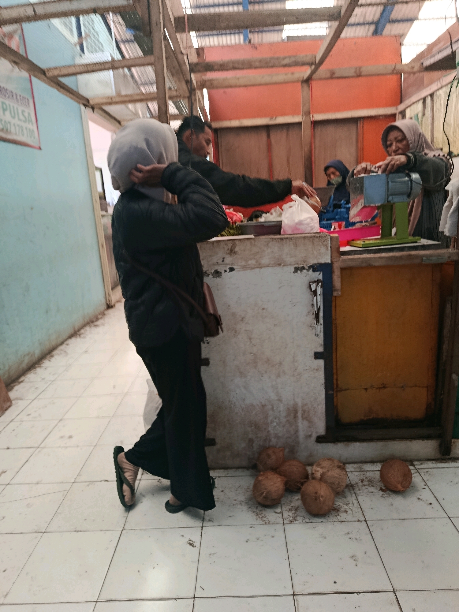 A group of people are gathered at what appears to be an outdoor market stall in Bululawang, Indonesia. In the foreground, a woman wearing a dark jacket and black pants, with a light-colored hijab, is walking away from the camera. She is carrying a brown bag slung over her shoulder. Nearby, several coconuts are scattered on the tiled floor. Behind her, a man in dark clothing is reaching towards a white plastic bag. To the right, two women are interacting with a food preparation machine. One woman, wearing a gray hijab and dark clothing, is operating the machine, while another woman, also wearing a hijab, is looking on. The stall is constructed with a corrugated metal roof supported by wooden beams. A blue and pink tray, along with other items, are visible on the stall. A sign with red and white text is mounted on a light blue wall in the background. The floor is tiled with a white checkerboard pattern.