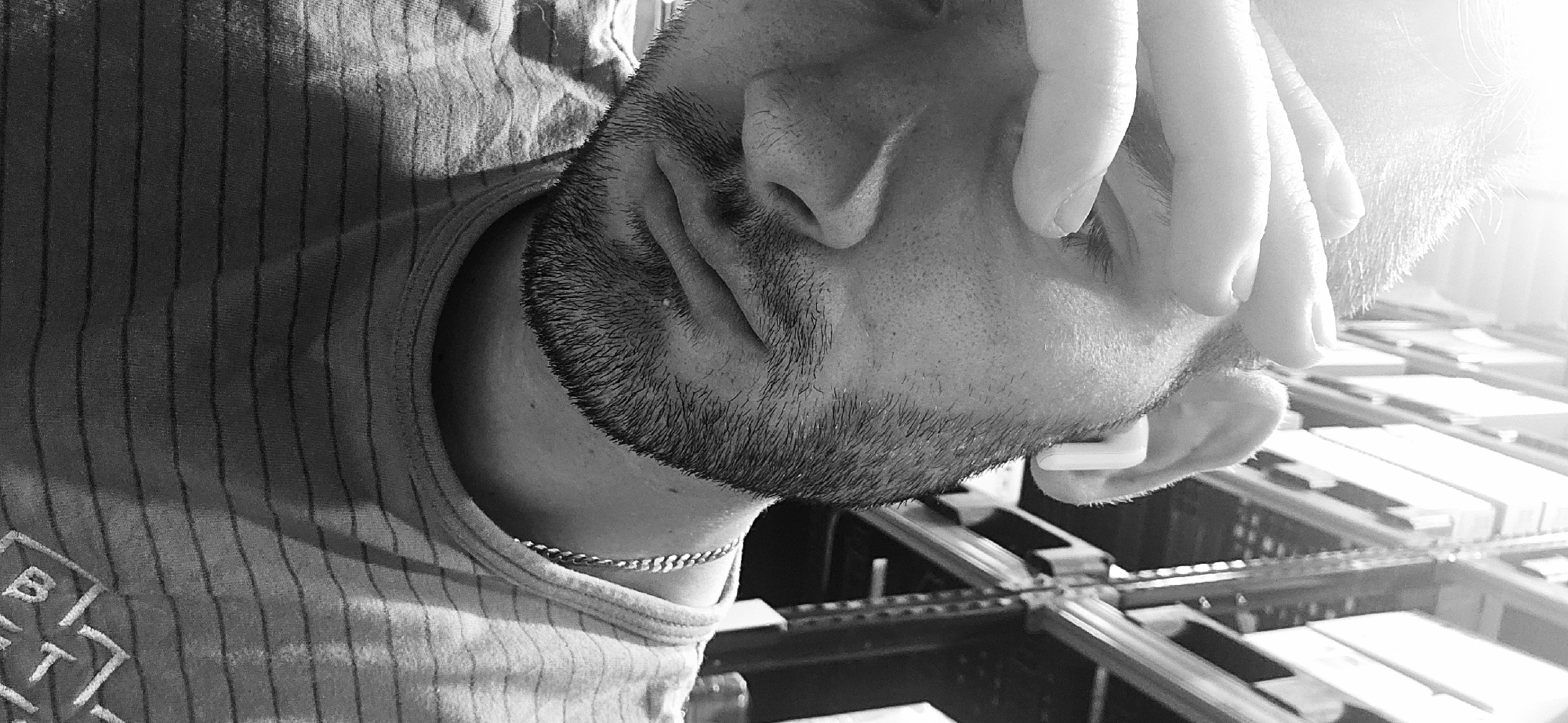 This black and white, close-up image, captured in Plovdiv, Bulgaria, features a person, likely male, depicted from the chest up, oriented sideways and looking downwards. The subject has short, dark stubble or a beard. Their right hand is raised, with fingers gently touching the side of their head or temple, suggesting contemplation or focus. A white earbud is visible in their ear, indicating they are listening to audio. Around their neck, a thin, light-colored chain necklace is present.
The person is wearing a light-colored, vertically striped sleeveless top or tank top. On the left side of the chest, a faint, light-colored logo or text is partially visible, appearing to show characters like "B" and "T" or "7". The background is blurred, showing rows of what appear to be shelves, racks, or machinery holding numerous rectangular objects, possibly suggesting an indoor environment such as an office, warehouse, or workshop. A strong light source from the upper right creates bright highlights and lens flare across the image.