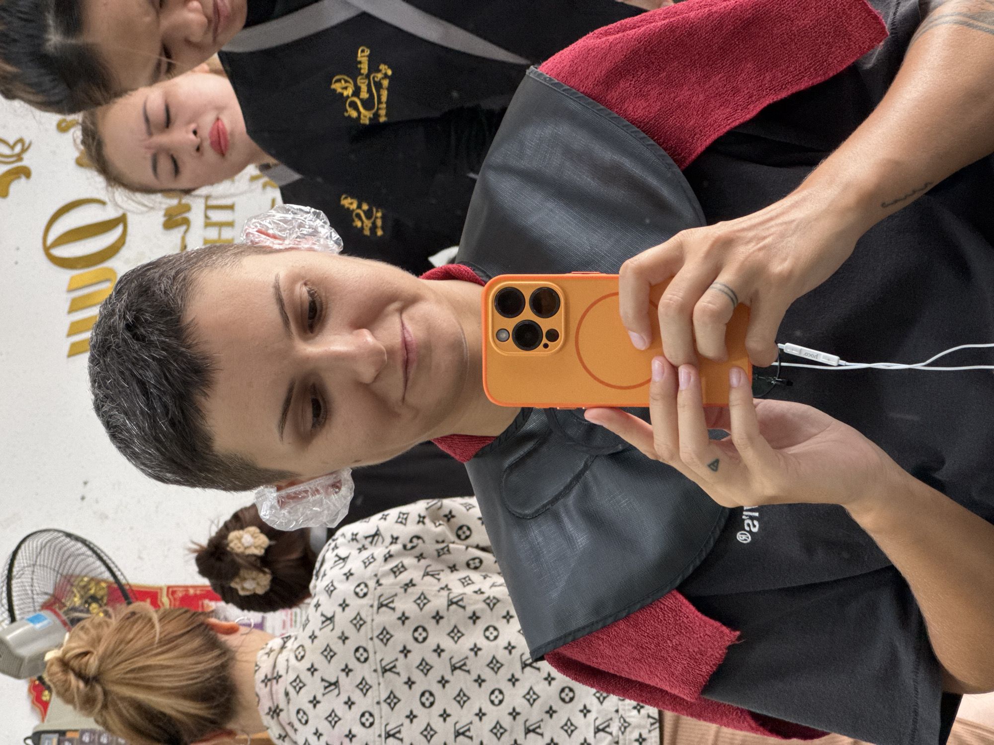 In a hair salon in Nha Trang Ward, Vietnam, a person with a short haircut and clear plastic coverings on their ears holds an orange smartphone in front of them, taking a selfie. The person is wearing a black t-shirt, a black cape over their shoulders, and a red towel draped around their neck. Two other individuals, appearing to be salon workers, are visible in the background. The person on the left has their hair pulled back in a bun and is wearing a white shirt with a repeating pattern. The person in the center has their eyes closed and is wearing a black uniform with a gold logo. The person on the right, also in a black uniform, is looking down. The salon environment includes a ceiling fan and a wall with signage.