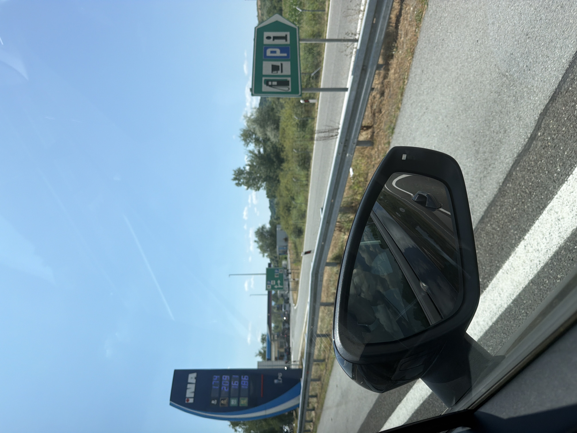 The image was taken from the interior of a vehicle traveling on a road in Općina Jakovlje, Croatia. In the foreground, a car's side mirror reflects the sky and surrounding landscape. To the left, a tall blue and white sign for the "INA" gas station displays fuel prices: 1.74, 2.09, 1.61, and 1.86. Beyond the sign, a gas station with multiple pumps and parked vehicles is visible. To the right, a green directional sign indicates services with icons for a gas pump, a cup of coffee, and an information symbol, pointing towards the right. The road is asphalt with white lines, and a metal guardrail runs alongside it. The environment is characterized by a clear blue sky with some faint contrails, green trees, and vegetation.