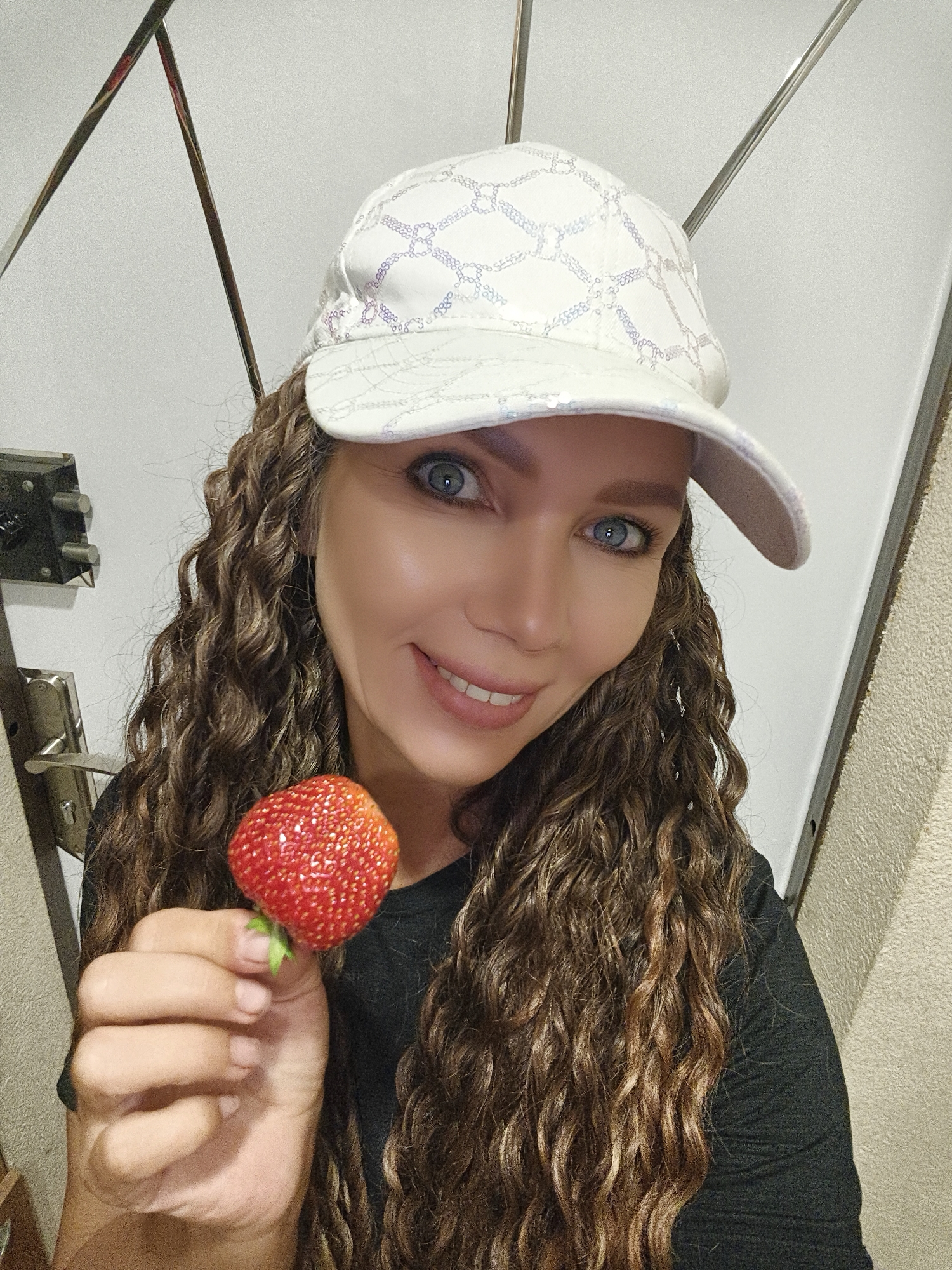 A woman wearing a white baseball cap and a black t-shirt holds a ripe strawberry in her right hand. She has long, curly brown hair and blue eyes. The background appears to be an indoor setting, possibly a hallway or entryway, with a wall that has some metallic elements and a door handle visible. There is no indication of the location being in the city of Chaikovsky, Russia.