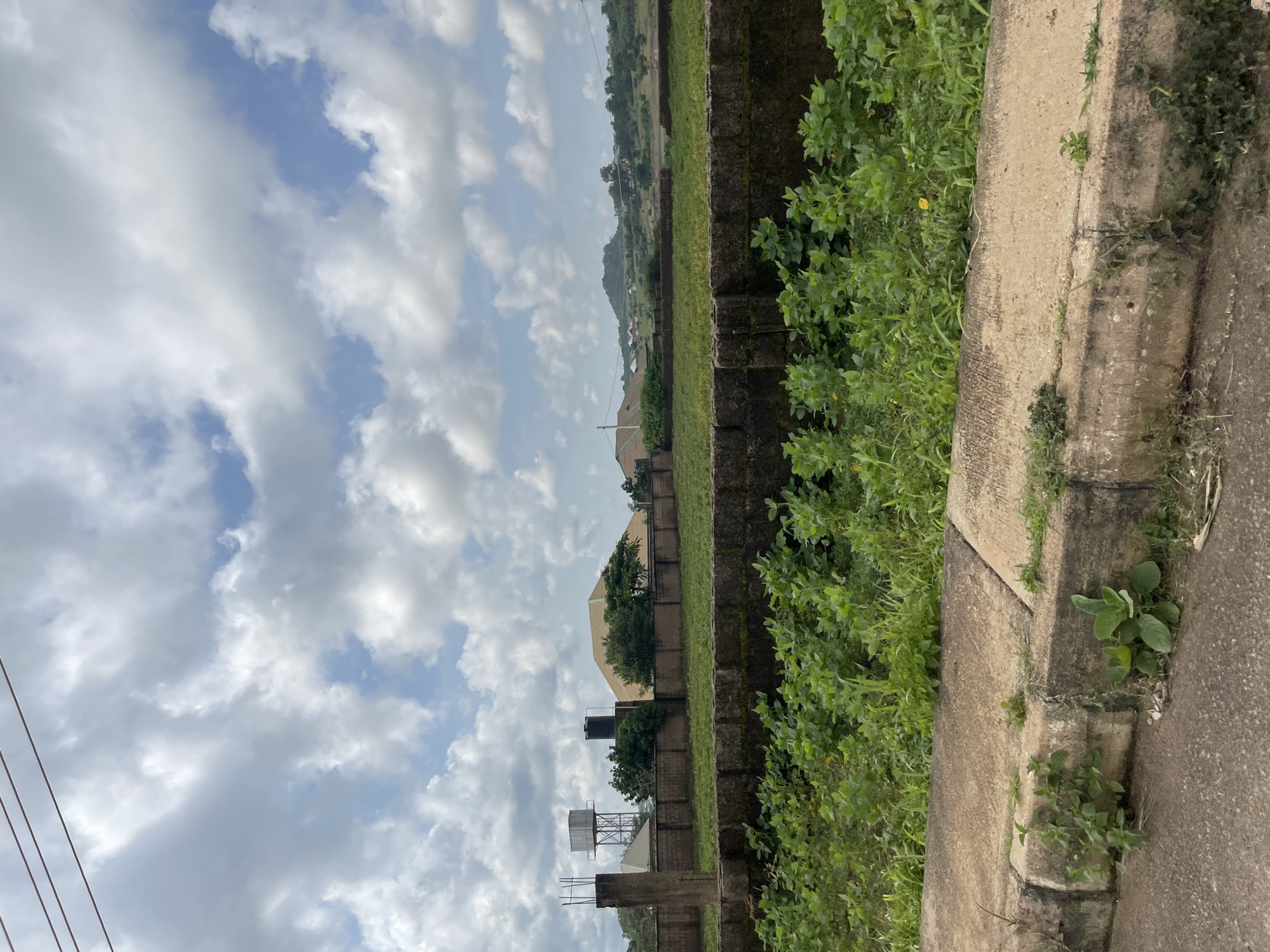 This is an outdoor, eye-level, wide shot image taken in Jos, Nigeria, on a cloudy day. The foreground consists of a concrete curb and a paved road. Behind the curb, a thick patch of green vegetation is visible, with some plants growing through the cracks in a low, dark-colored stone wall.
Beyond the vegetation and wall, there is a large, empty plot of land that appears to be undergoing some form of development or is intended for future construction. A concrete pillar, possibly a remnant of a foundation or fence, stands prominently on the left side of the plot. In the background, a tall metal water tower with a tank on top is visible, along with another black water tank. Further back, there are several buildings with distinctive roofs, one with a pale yellow, curved roof. The landscape gently slopes upwards towards a distant hill. The sky is filled with a mix of white and gray clouds, with patches of blue sky showing through. The overall impression is one of an open space within an urban or semi-urban environment.