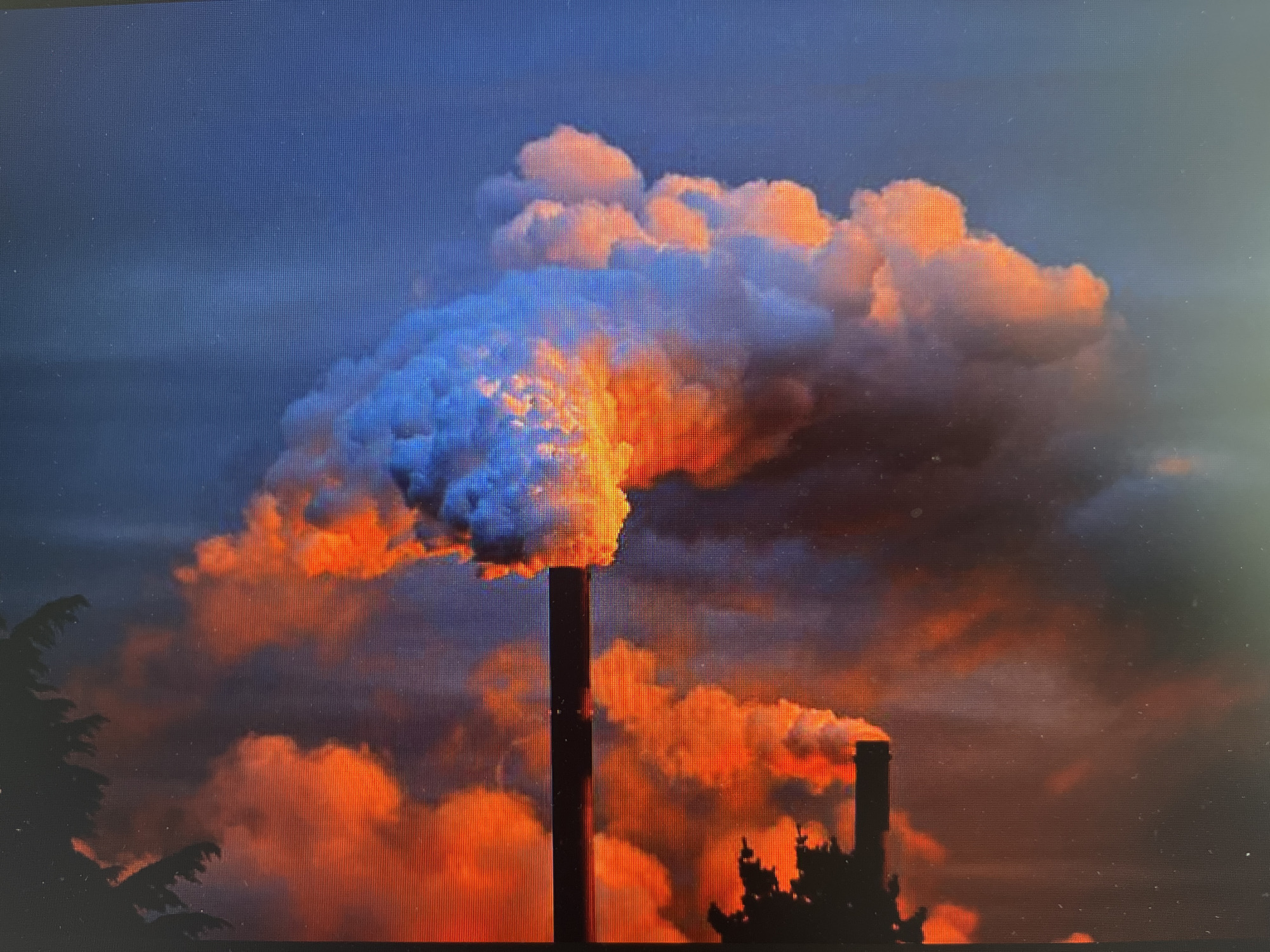 The media file presents an industrial landscape featuring two prominent smokestacks actively emitting large plumes of smoke or steam. The central, taller smokestack is dark and vertical, releasing a dense cloud that billows upwards and outwards. This cloud is intensely illuminated with vibrant orange and red tones, particularly where it emanates from the stack, contrasting with cooler blue and grey hues along its edges and in the surrounding atmosphere. A second, smaller smokestack is visible towards the right, also contributing to the emissions with a reddish-orange plume.
The sky exhibits a dramatic color palette, transitioning from deep blues and dark greys in the upper regions to warmer orange and red tones that intermingle with the industrial emissions. This striking coloration strongly suggests either sunrise or sunset. Silhouetted dark trees or foliage are visible on the far left and in the lower right, partially framing the industrial structures. The overall scene depicts a powerful visual contrast between the stark, man-made structures and the vivid, illuminated natural sky, heavily influenced by the industrial output.
The image itself appears to be a photograph of a digital screen or display, as evidenced by the visible grid-like pixelation across the entire frame and minor dust specks. No discernible text is present within the image. While the location is Varna, Bulgaria, no specific geographical cues within the image confirm this.