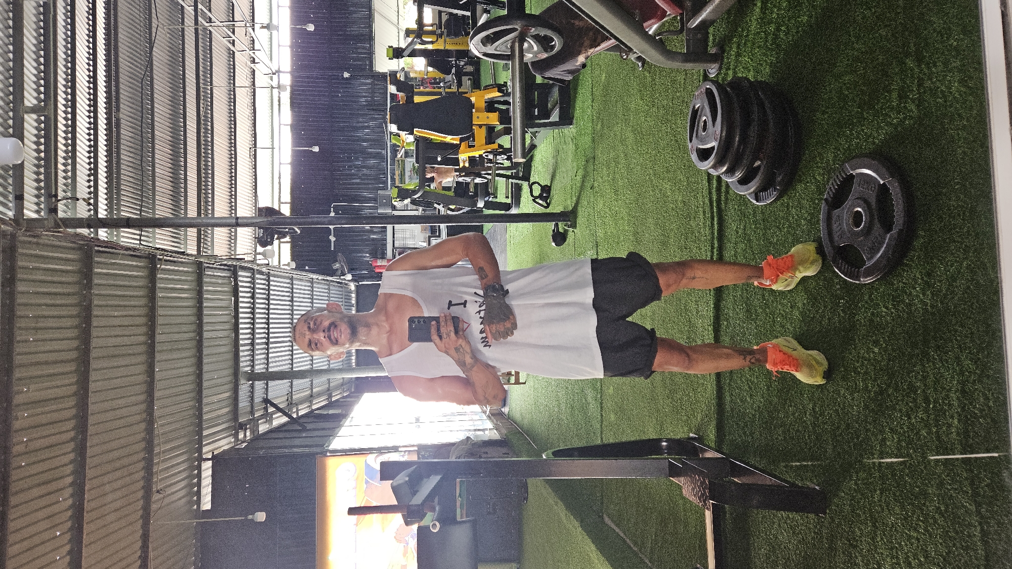A man in a tank top and shorts stands in a gym, holding a phone and looking at the camera. The gym is located in Cam Ranh Ward, Vietnam. Weightlifting equipment and weights are visible. The floor is covered in artificial turf. The gym has a corrugated metal roof.