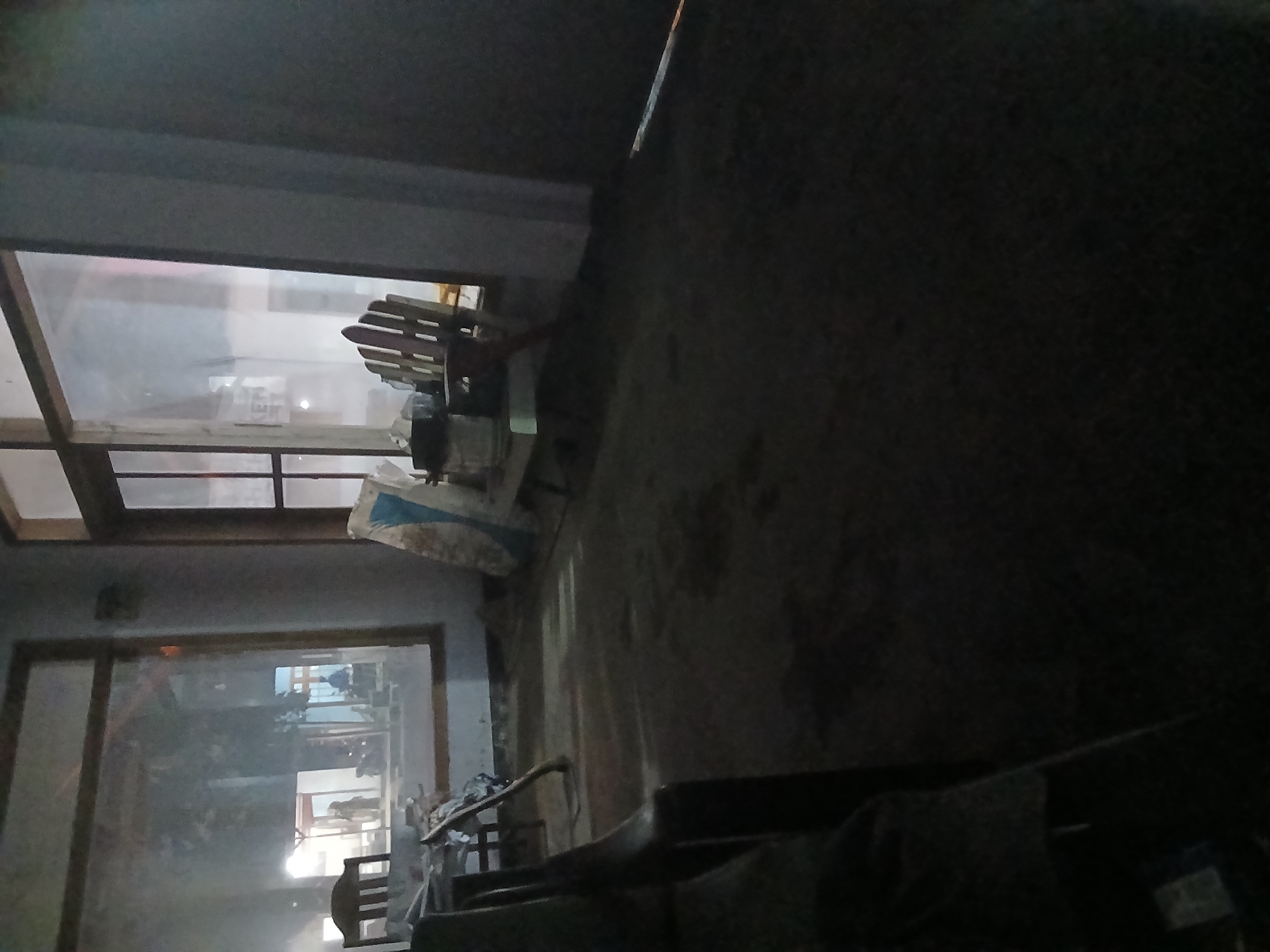 The image depicts a dimly lit indoor scene, likely a room or a covered outdoor area. Two wooden chairs are visible, one on the left with a dark seat and back, and another on the right, a slatted Adirondack-style chair. In the background, through large windows or openings, a blurry scene of what appears to be a street or public area is visible with reflections. There are indistinct shapes that could be people and vehicles in the background. Several objects are scattered on the floor and near the chairs, including a white bag with blue markings, possibly a sack of cement or similar material, and other unidentifiable items. The floor is uneven and appears to be dirt or loose debris. The overall impression is one of disarray or an unfinished space. The location in Kademangan Kulon, Indonesia, suggests a tropical or sub-tropical environment, but the image itself does not provide specific visual cues to confirm this.