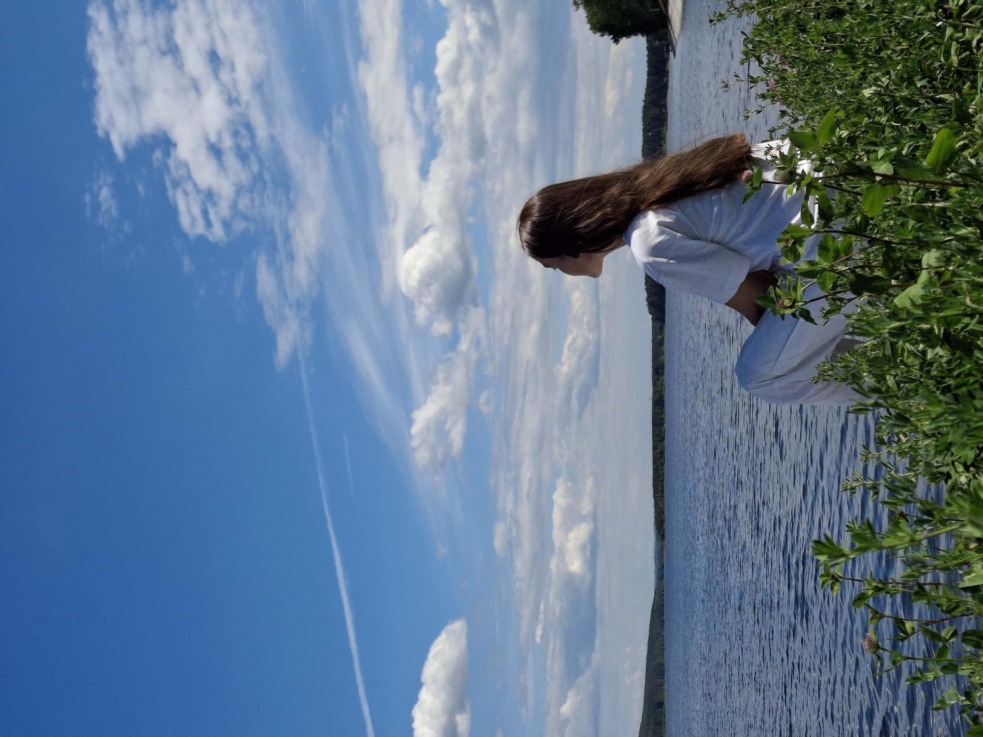 The image depicts a lone female figure seated by the Votkinsk Reservoir on the Kama River, near Chaikovsky, Russia. The woman is viewed from behind and slightly to her right, with her head turned to the left, gazing towards the horizon over the expansive body of water. She has long, straight, dark brown hair extending down her back and is wearing a loose-fitting white t-shirt and light-colored trousers. A dark earbud or small headphone is visible in her left ear. The expansive body of water exhibits a blue hue with a subtly rippled surface, suggesting a slight breeze. A dark green forested shoreline stretches across the distant horizon. The sky above is predominantly clear blue, interspersed with scattered white cumulus clouds, particularly dense in the mid-left and upper-right areas. A prominent white contrail from an aircraft traverses the upper left portion of the sky, extending diagonally downwards. The immediate foreground features lush green vegetation, including broadleaf plants and some small pinkish-purple flowers, partially obscuring the lower part of the woman's body.