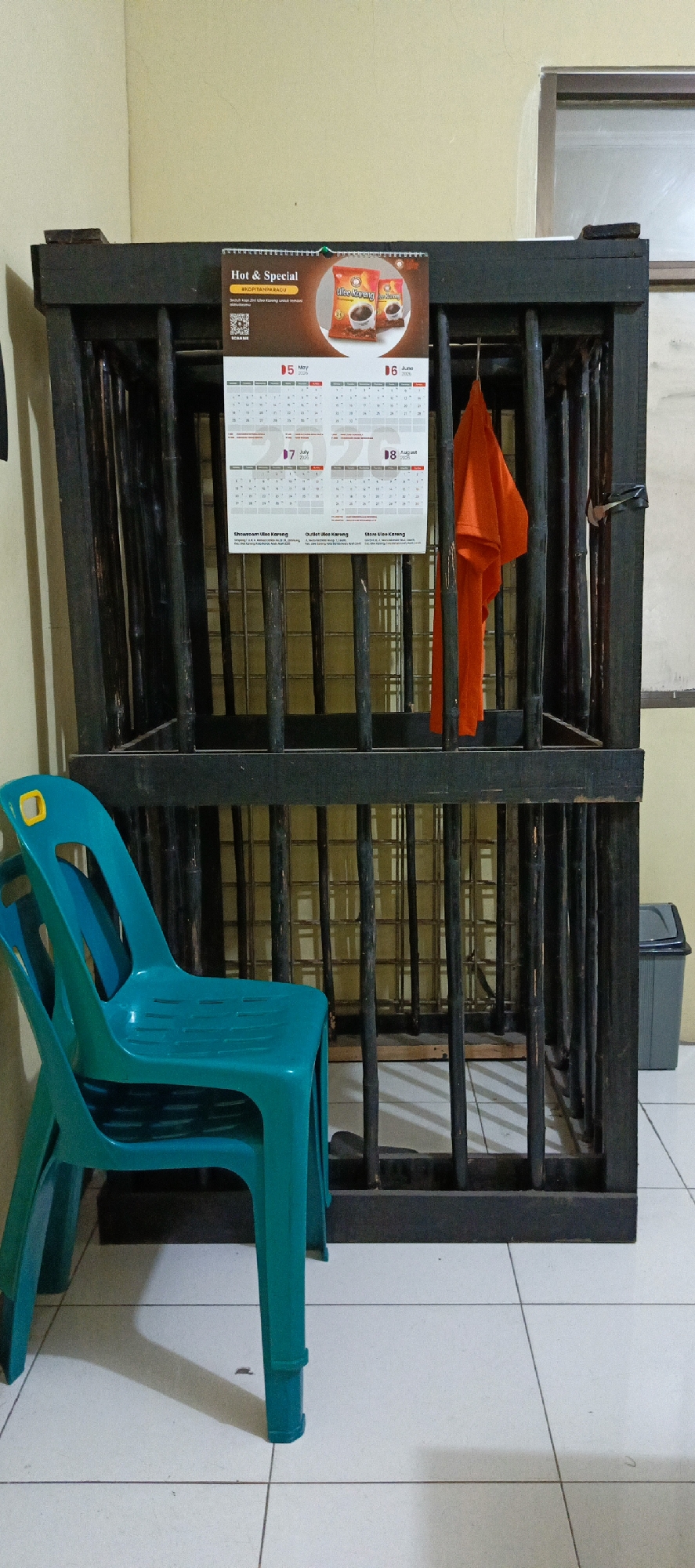 The image depicts an indoor scene featuring a dark, wooden cage-like structure with vertical bars, situated on a tiled floor. A teal plastic chair is positioned in front of the cage. Hanging inside the cage is a bright orange piece of fabric. A calendar is attached to the top of the cage, displaying months and dates. To the right of the cage, a small gray trash bin is visible. The walls appear to be a light yellow color. The overall impression is that of a utilitarian or storage space, possibly in a residential or commercial setting in Banda Aceh, Indonesia.