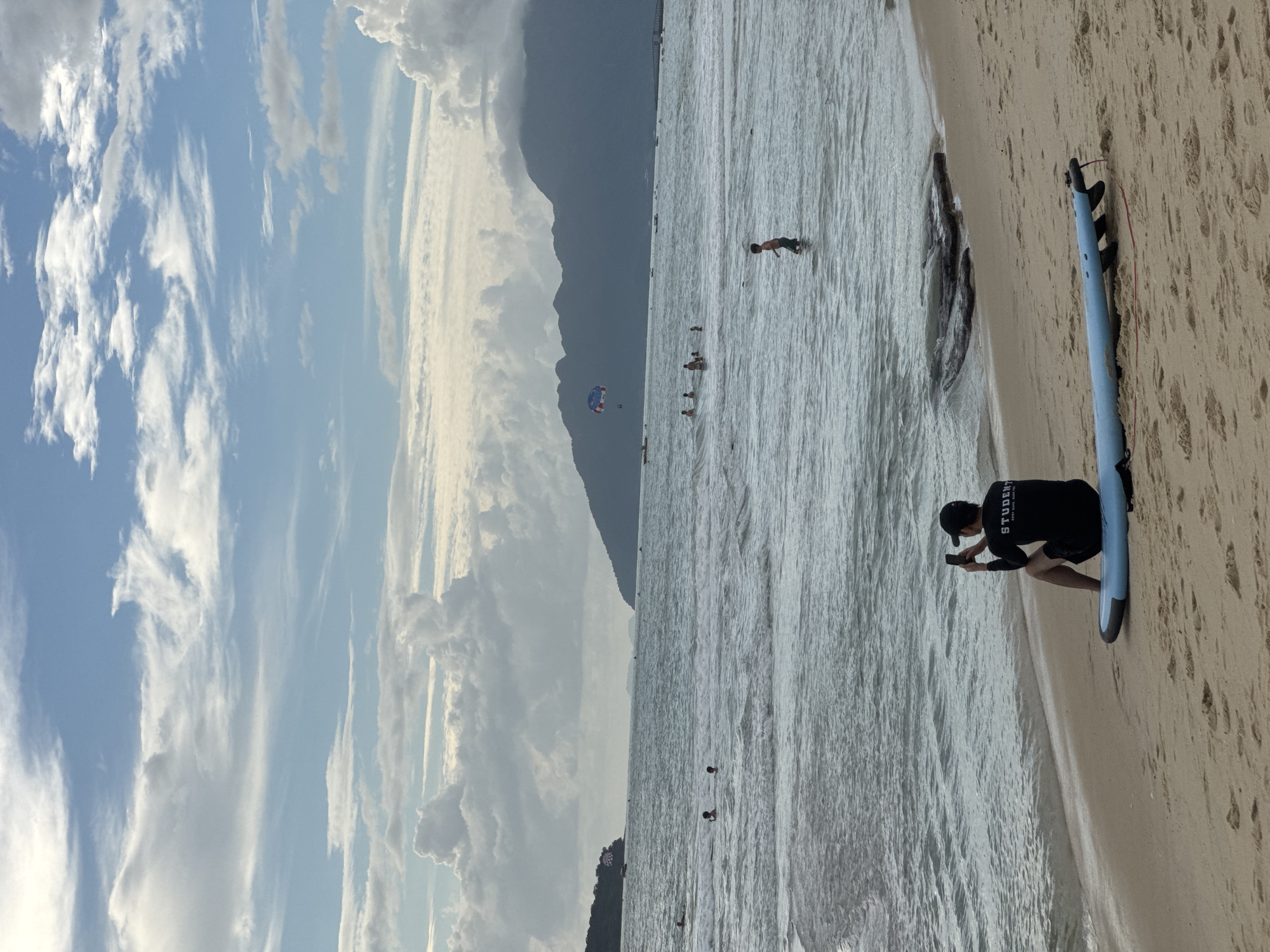 The image depicts a beach scene on Tengah Island, Malaysia. In the foreground, a person wearing a black t-shirt with the word "STUDENT" on it and black shorts sits on a blue surfboard, facing away from the camera, holding a mobile phone up as if taking a picture. The beach is sandy, with some footprints visible. The ocean is to the left, with gentle waves reaching the shore. In the water, several other individuals are visible, some swimming or wading. Further out in the sea, a parasail is being towed by a boat, and mountains form a backdrop to the horizon under a cloudy sky.