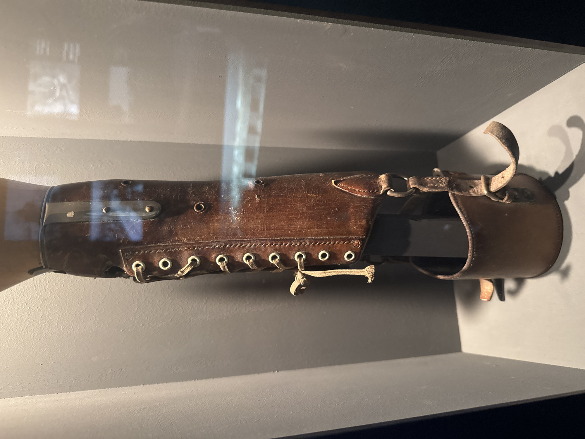 The image shows a close-up, side view of an antique leather prosthetic forearm. It is displayed in a museum setting, likely in a glass case as evidenced by reflections. The prosthetic is made of dark brown leather with visible wear and tear, suggesting age and historical use. It features a lacing system with metal eyelets and a fabric cord, similar to a boot, for adjustment and attachment. There are also metal studs or rivets along the length of the prosthetic, as well as a buckle and strap mechanism at the upper end. The prosthetic is mounted on a mannequin or a base, simulating it being worn. The background is a muted, neutral gray, which allows the prosthetic to be the focal point. Reflections on the glass reveal faint outlines of what appear to be exhibits or architectural features, hinting at the museum's interior. The lighting is somewhat dim, creating shadows that add to the artifact's historical feel. The overall impression is of a preserved piece of medical history, possibly from the late 19th or early 20th century, presented for educational purposes. Given the location context of Bern, Switzerland, this artifact could be part of a collection at a Swiss museum detailing medical history or the history of disability aids. There is no visible text in the image.