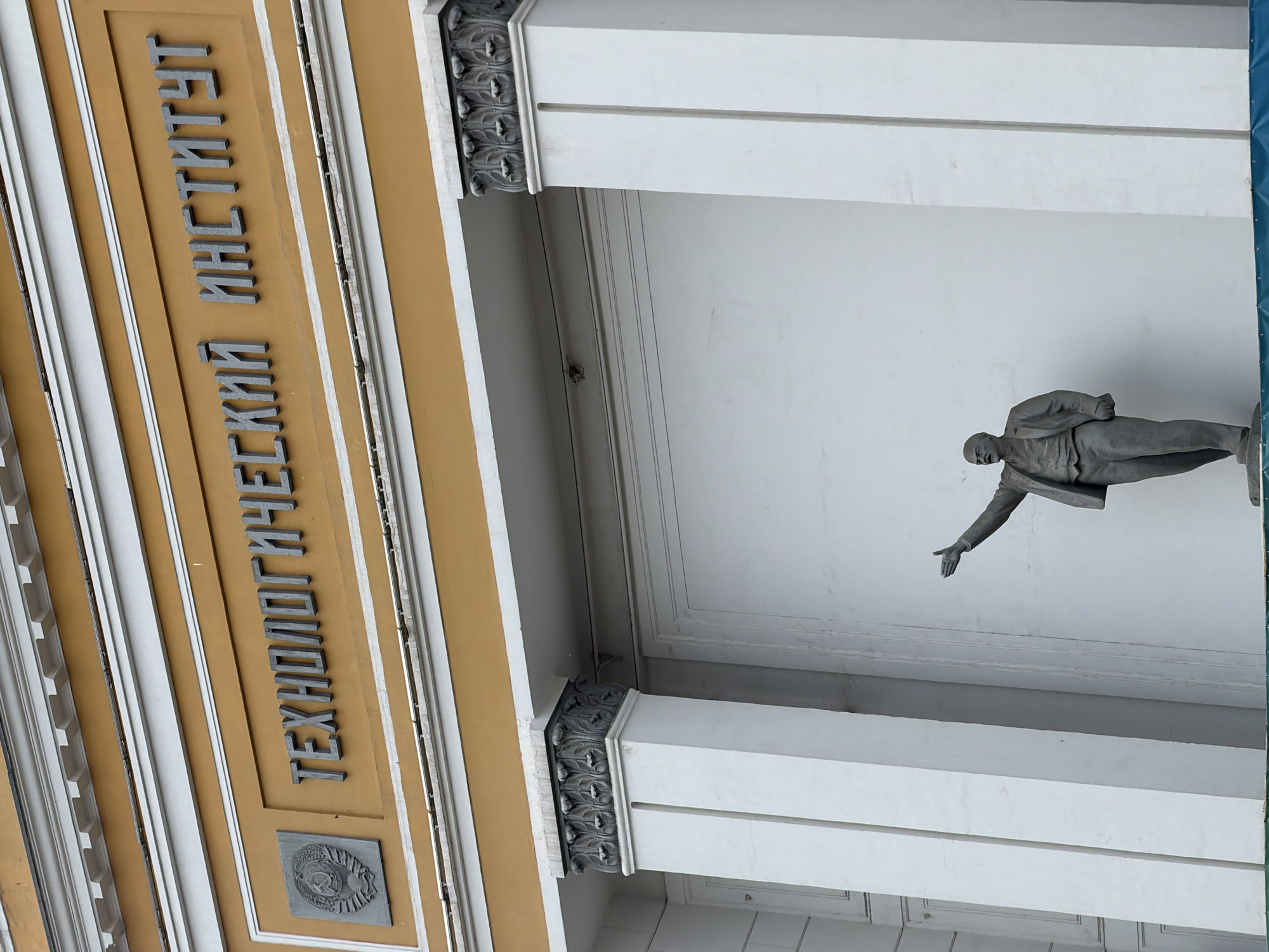 A low-angle, close-up shot shows the facade of a building with the inscription "ТЕХНОЛОГИЧЕСКИЙ ИНСТИТУТ" (Technological Institute) in large, grey, three-dimensional letters. To the left of the inscription is a grey, square emblem with a hammer and sickle. The building features white columns with decorative grey capitals and an orange-colored band above the inscription. In the foreground, a grey statue of a man in a suit, identified as Vladimir Lenin, stands with his right arm outstretched and pointing. The statue is positioned against a white wall. The overall scene appears to be an exterior view of an educational institution in Krasnoyarsk, Russia, likely the Krasnoyarsk State Technological University, based on the inscription.